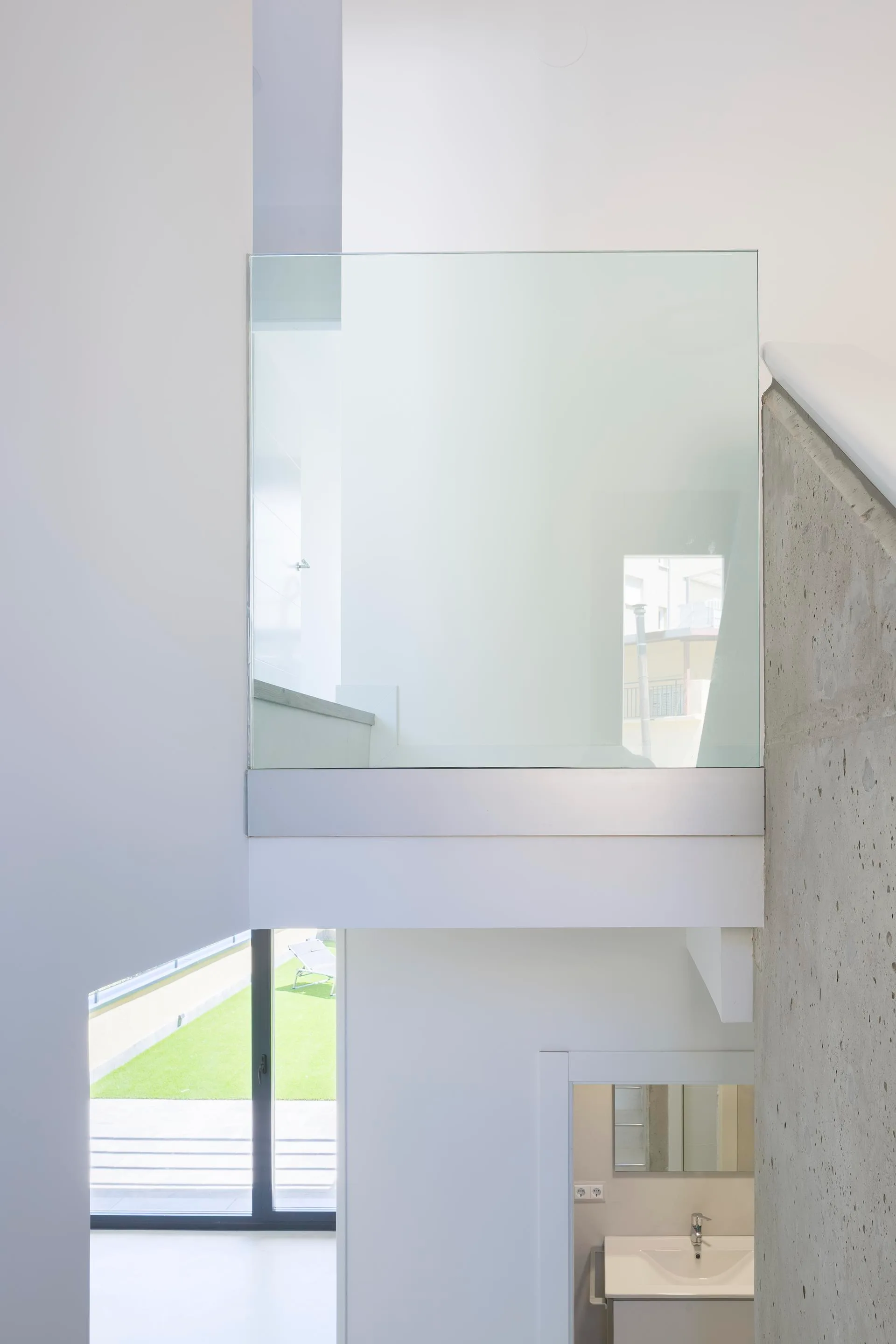 A minimalist interior view shows a staircase with glass railing panels next to a textured concrete wall, with glimpses of a bathroom and garden below.
