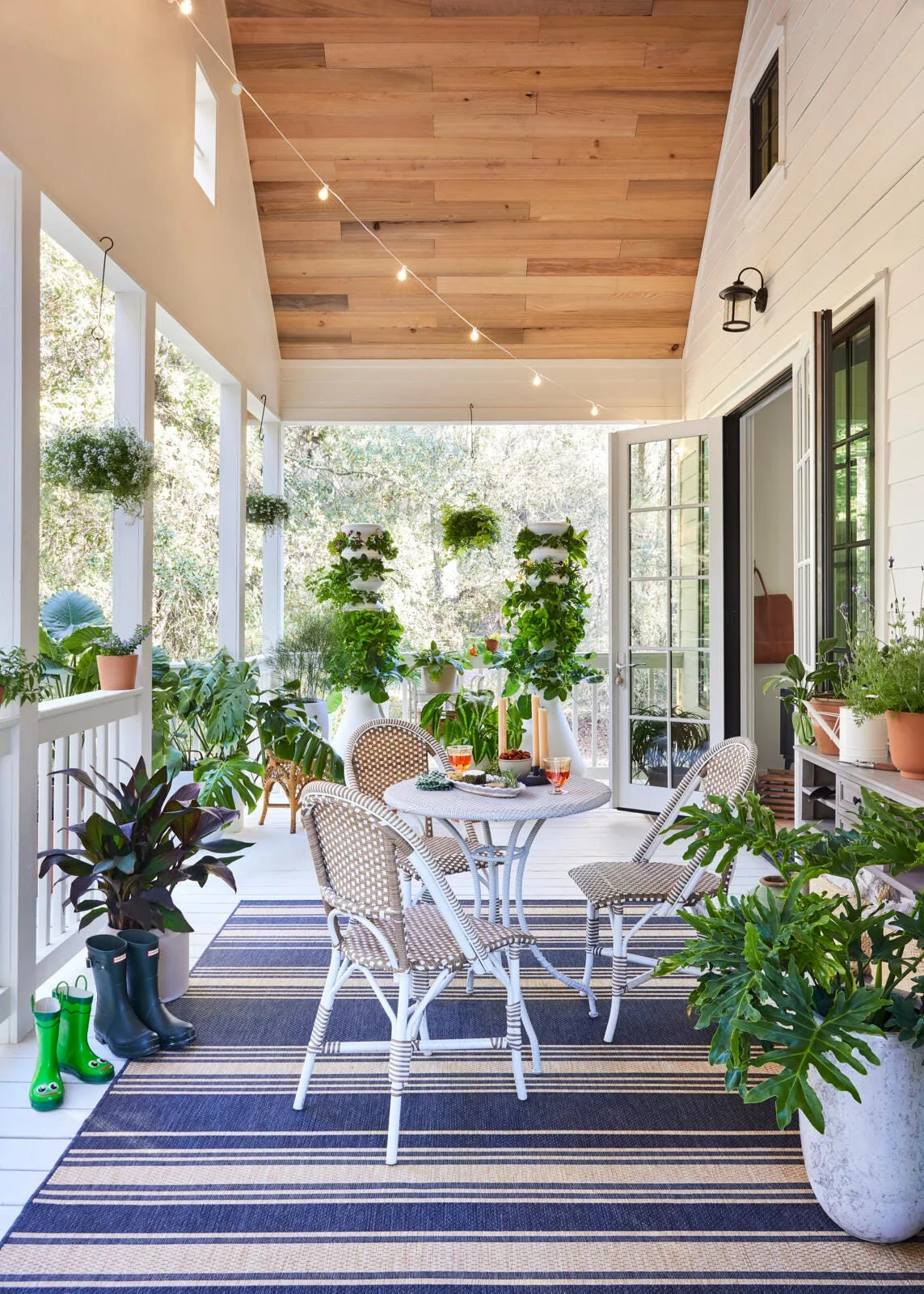 A covered porch featuring a woven bistro dining set on a striped blue and white rug, surrounded by potted plants under a wood-paneled ceiling.