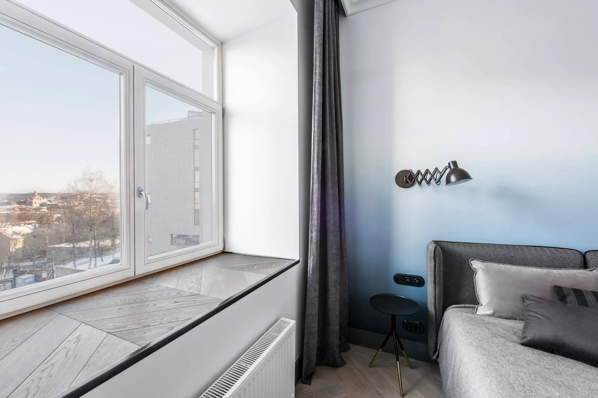 A modern bedroom corner features a blue gradient wall, a gray bed with textured throw pillows, a black wall lamp, and a herringbone-patterned window sill.