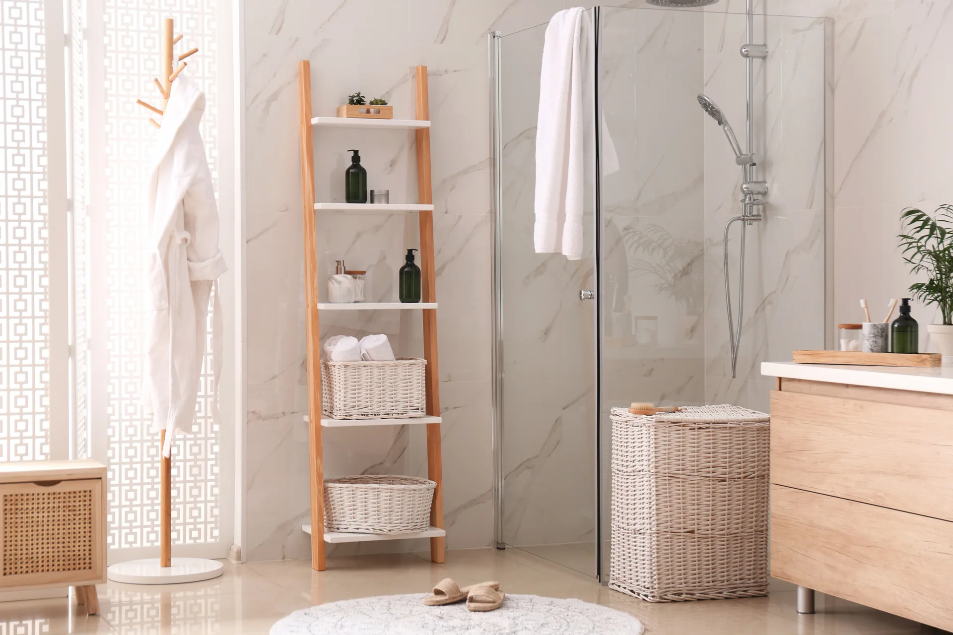 A bright bathroom features a light wood vanity, a glass-enclosed shower with marble tiles, a wooden ladder shelf, and white wicker baskets.