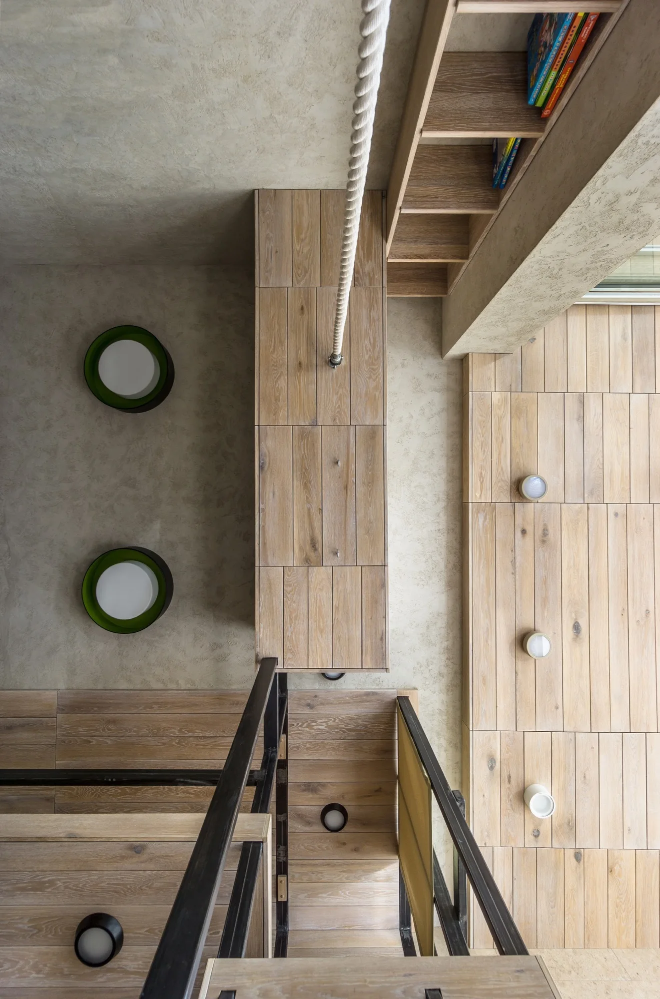 An overhead view of a modern wooden staircase featuring a black metal handrail, textured beige plaster walls, and round wall sconces.