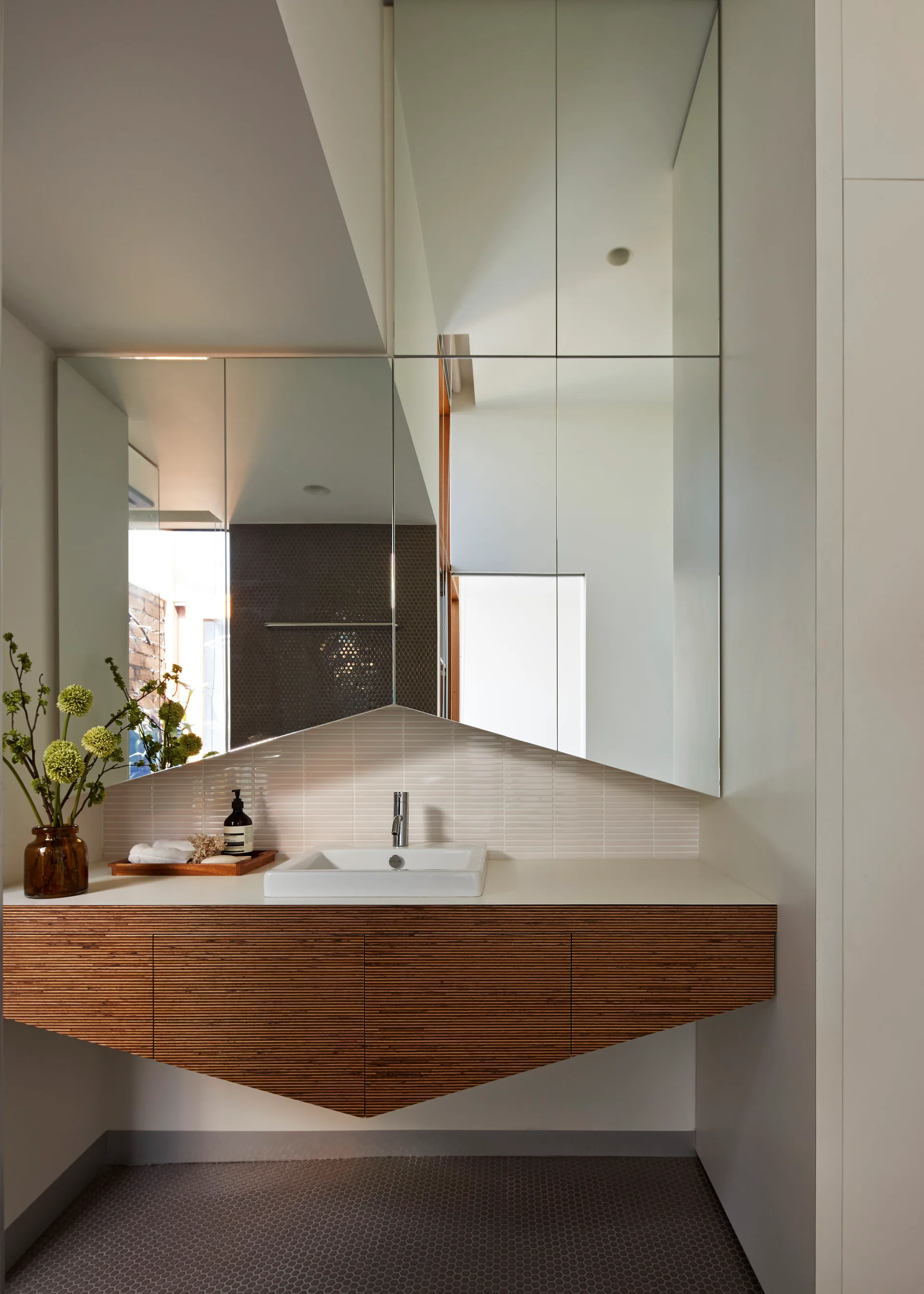 A floating slatted wood vanity is paired with an angular geometric mirror, a white sink, and a tiled backsplash in a modern bathroom.