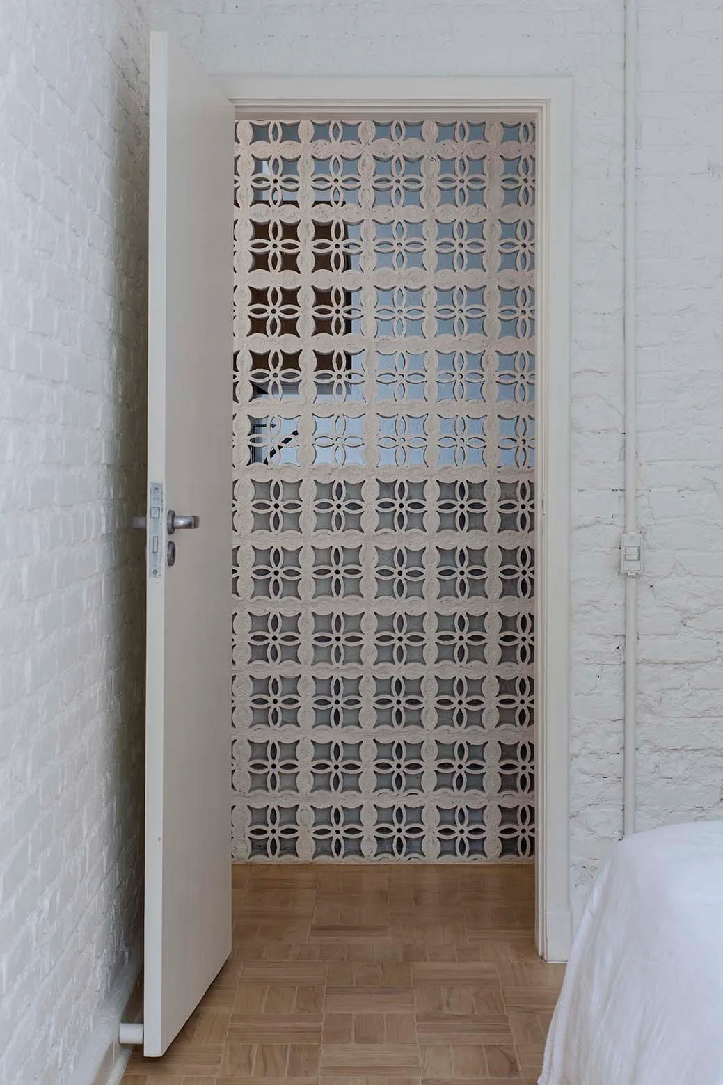 An open white door reveals a geometric concrete breeze block screen framed by painted white brick walls and light wood parquet flooring.