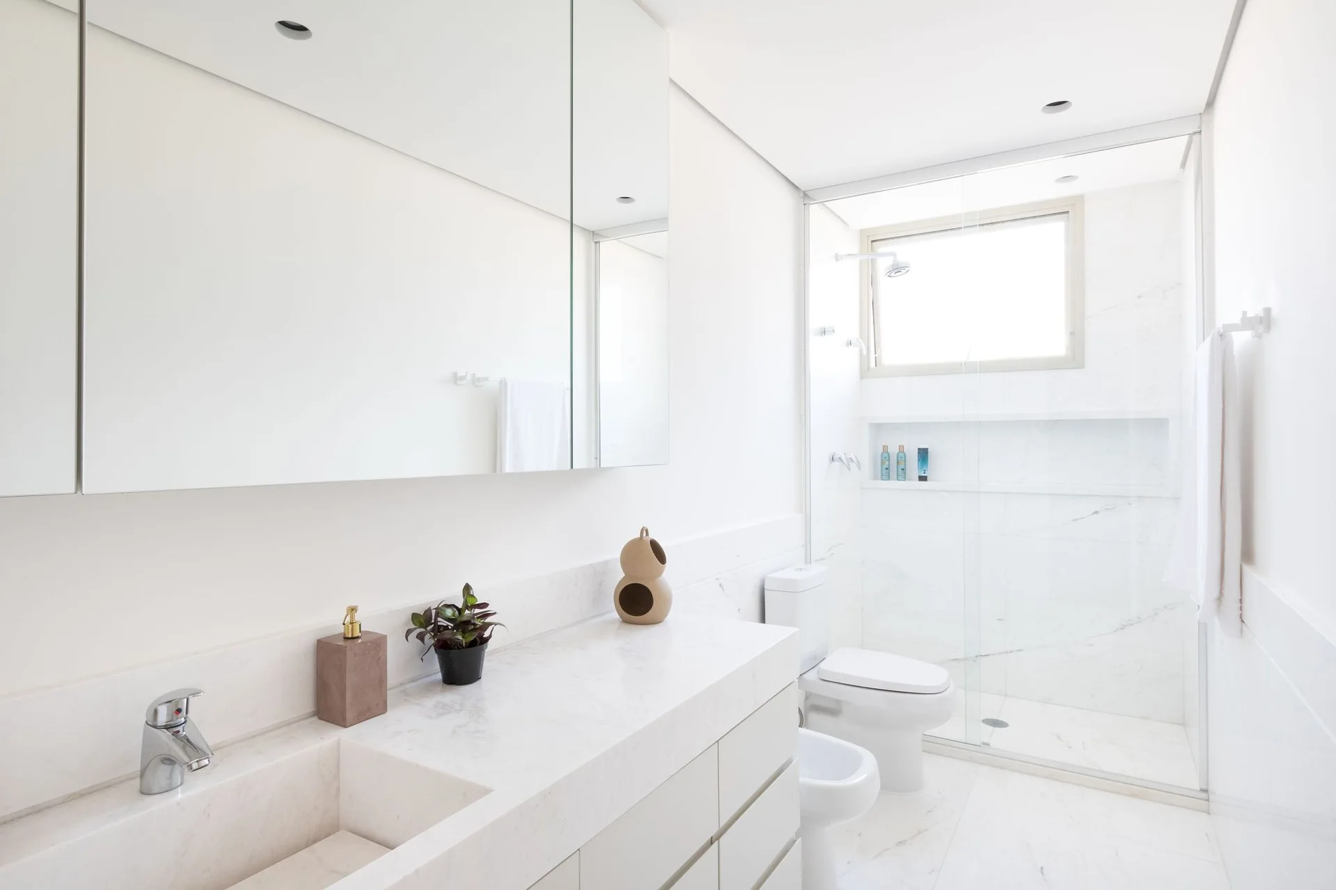 A minimalist white bathroom featuring a marble vanity with an integrated sink, a large frameless mirror, and a glass-enclosed shower.