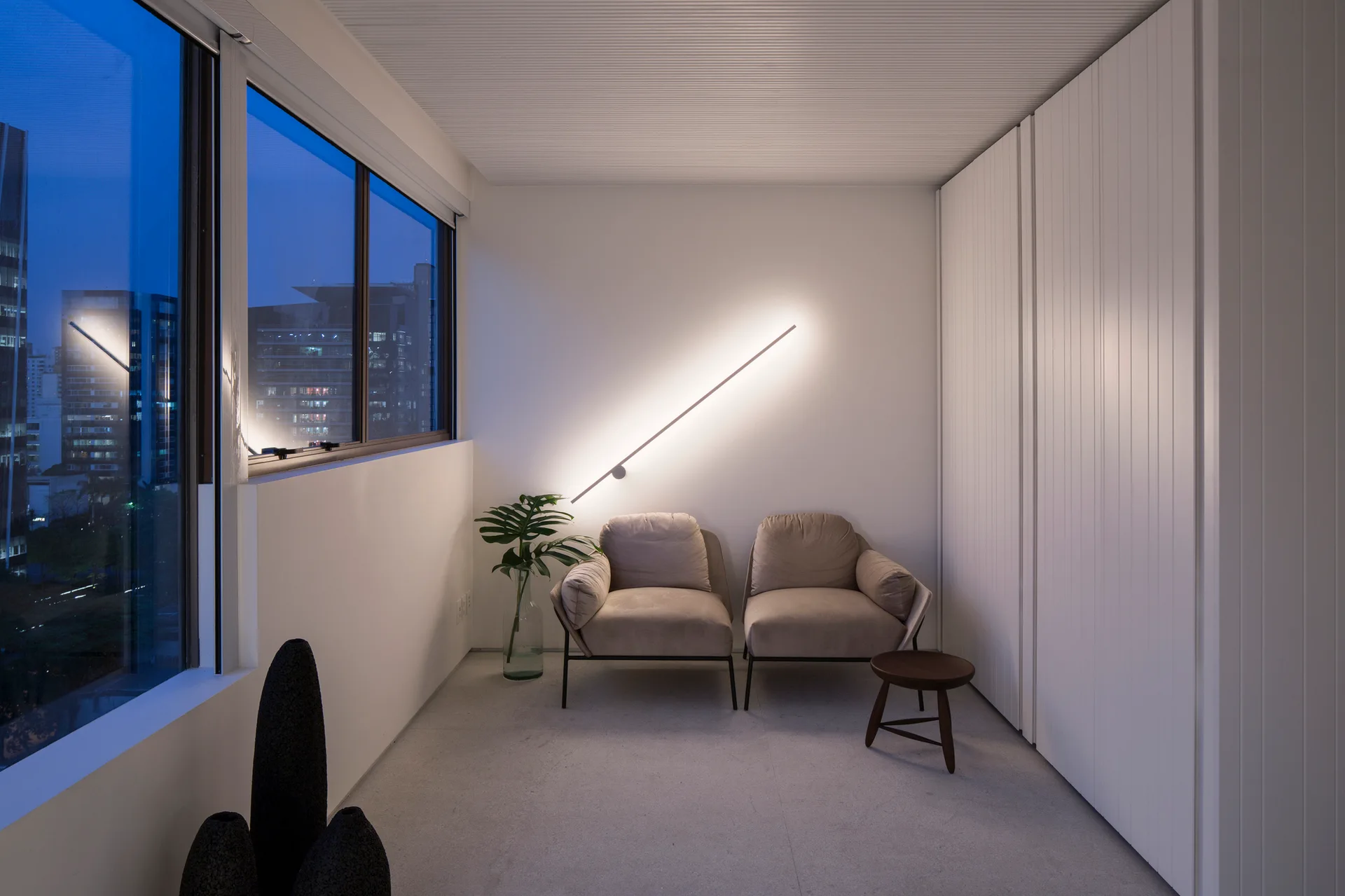 Two beige armchairs stand next to a large window under a glowing diagonal light strip mounted on a plain white wall.