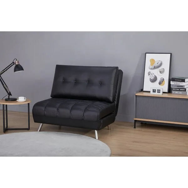 A black tufted sofa bed rests on wood flooring next to a round side table with a lamp and a slatted console cabinet against a grey wall.
