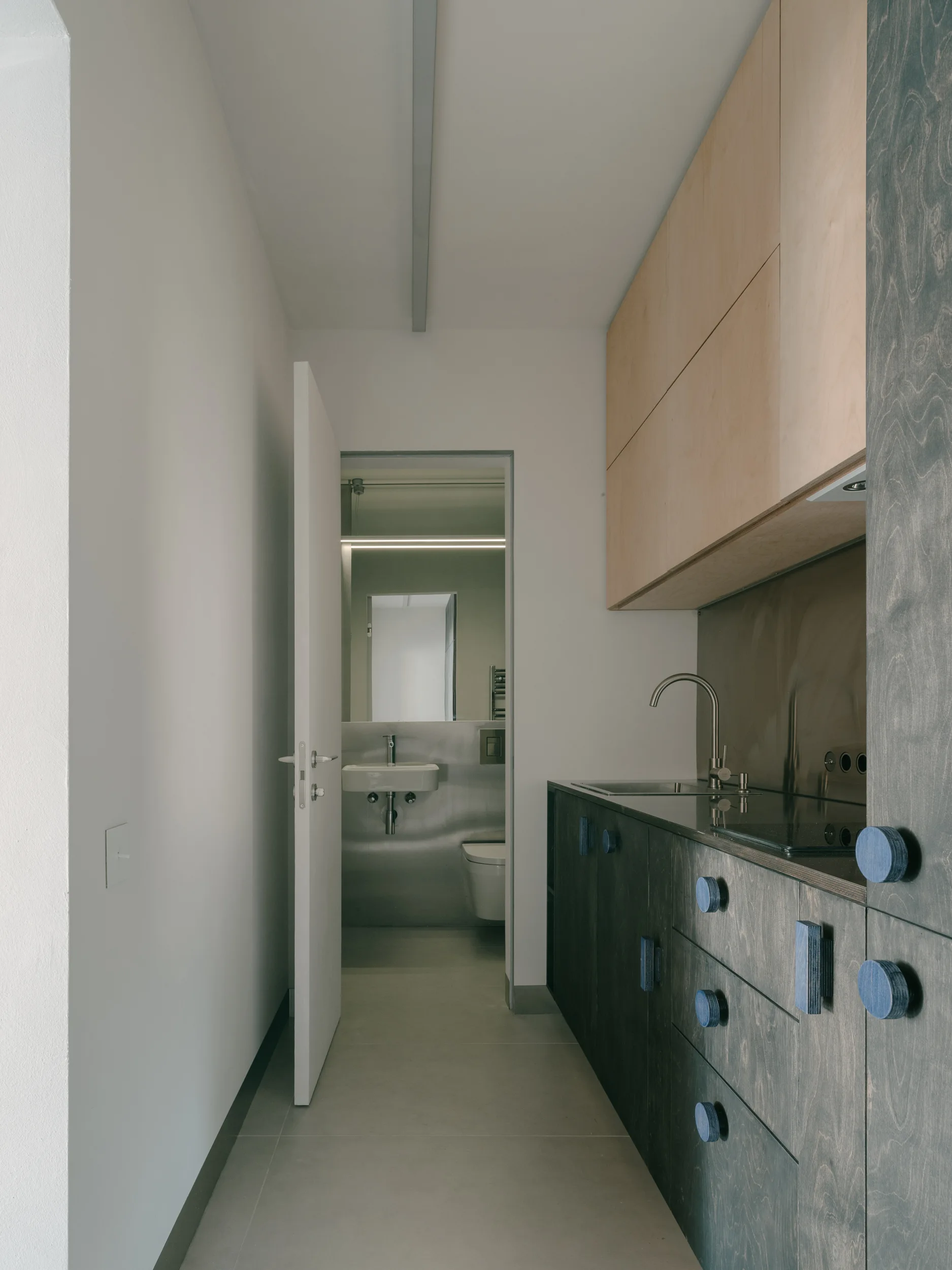 A narrow galley kitchen features dark grey lower cabinets with blue circular knobs, light wood upper cabinets, and an open doorway to a grey tiled bathroom.