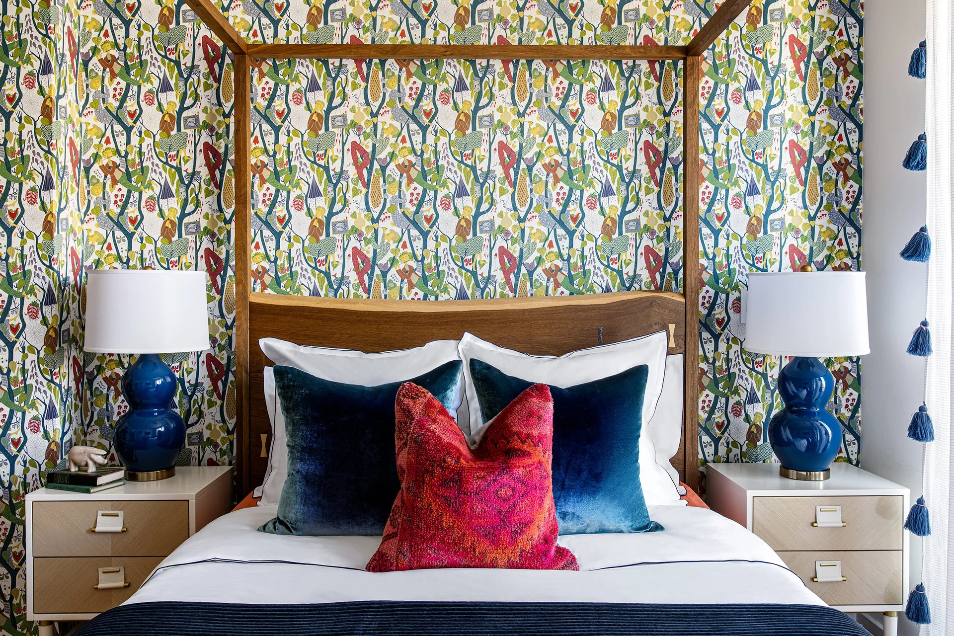 A wooden canopy bed with blue and red pillows sits against colorful botanical wallpaper, flanked by nightstands with glossy blue gourd lamps.