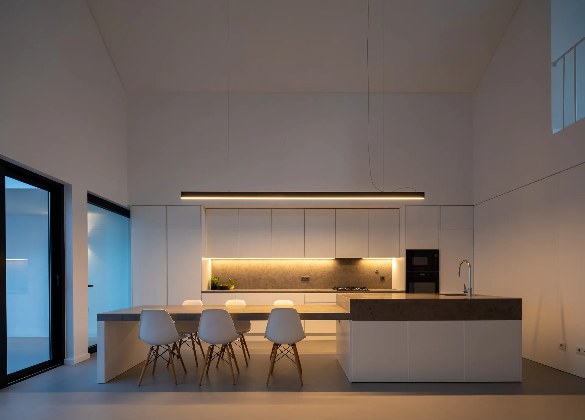 A minimalist kitchen features matte white handleless cabinets, a gray stone island integrated with a dining table, and a long black linear pendant light.