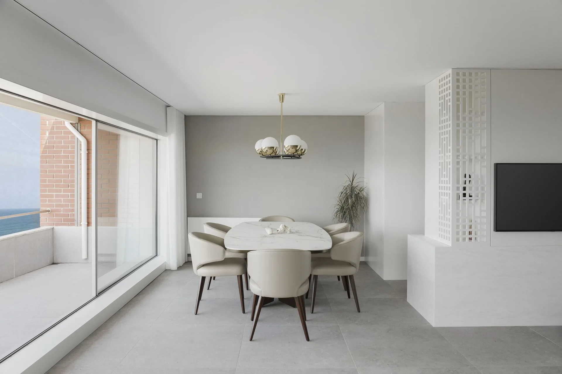 A modern dining room features an oval marble-top table with six beige chairs beneath a globe pendant light, next to a large sliding glass door.