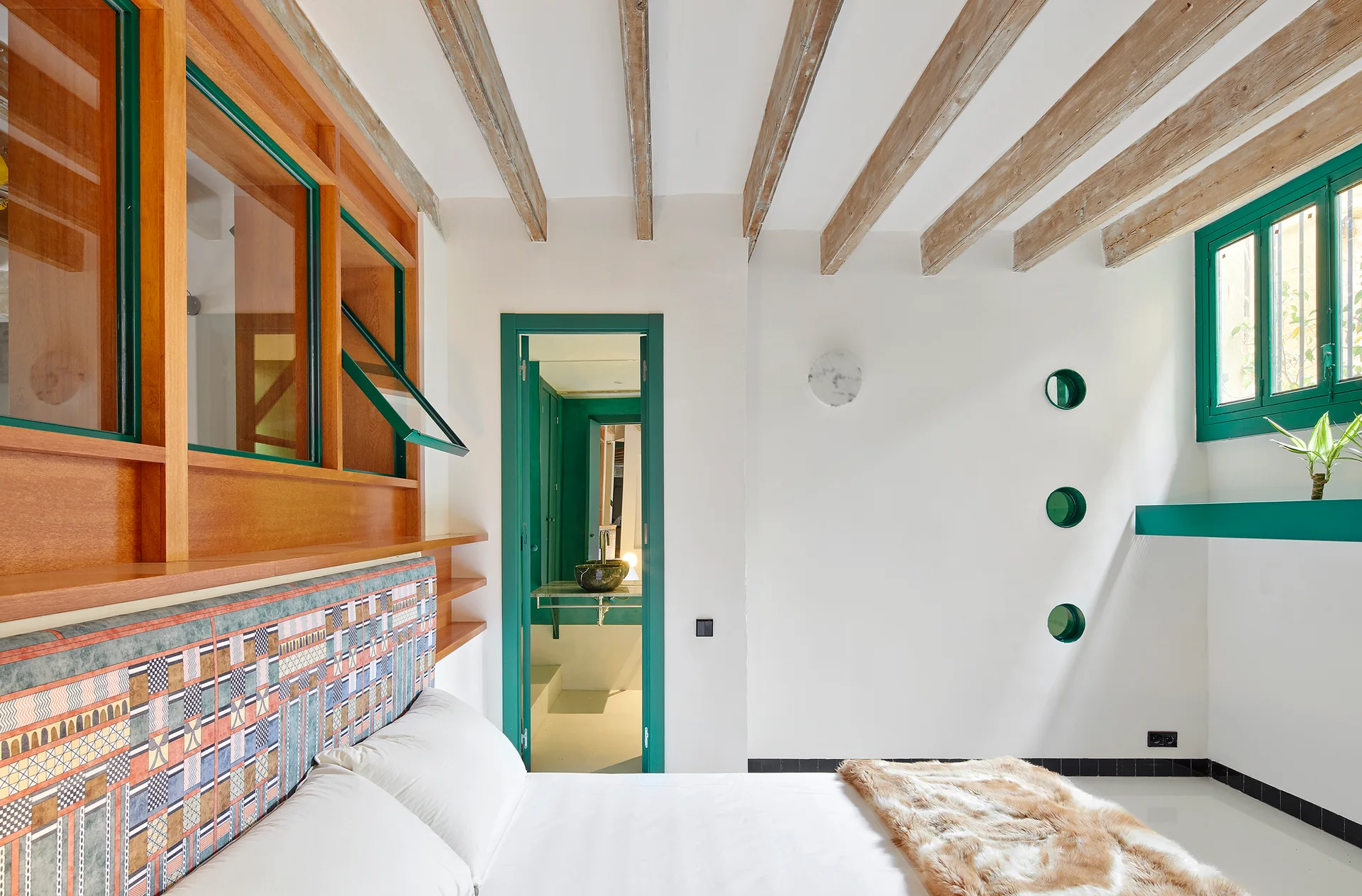 A bright bedroom features a bed with a patterned fabric headboard, exposed wood ceiling beams, and door and window frames painted deep green.