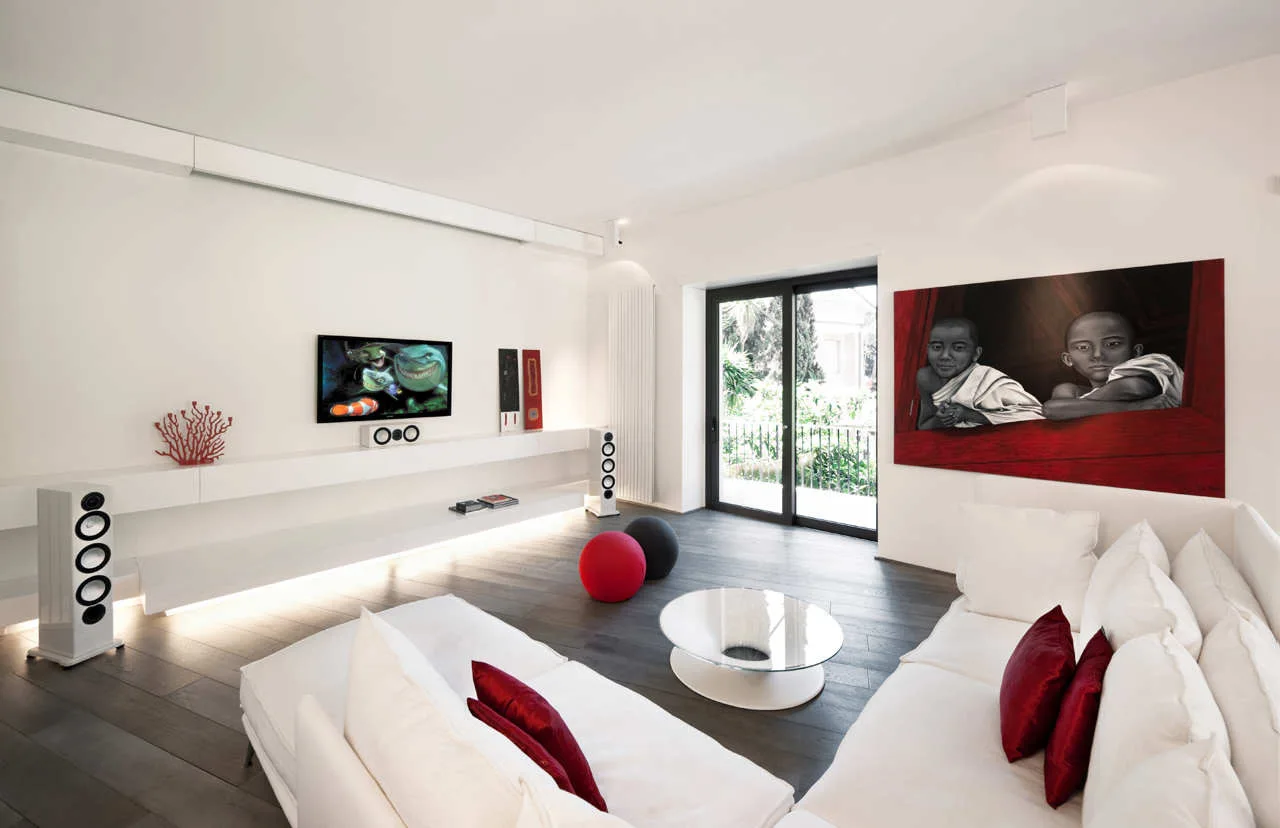A white minimalist living room features a long floating wall console with integrated under-lighting, a white sectional sofa, and a large red-accented painting.