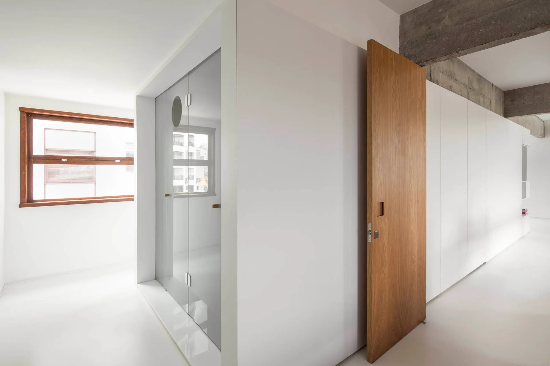 A minimalist corridor features integrated flat-panel white closets, a warm wood door, and an exposed concrete ceiling beam.