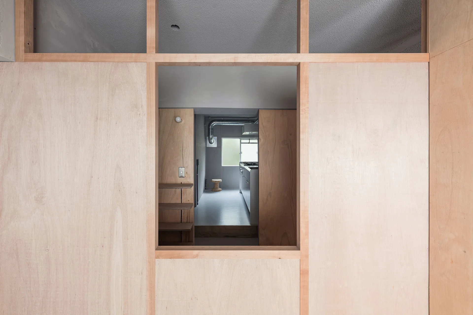 A light-toned plywood partition wall features a large rectangular cutout looking into a minimalist kitchen with gray floors.