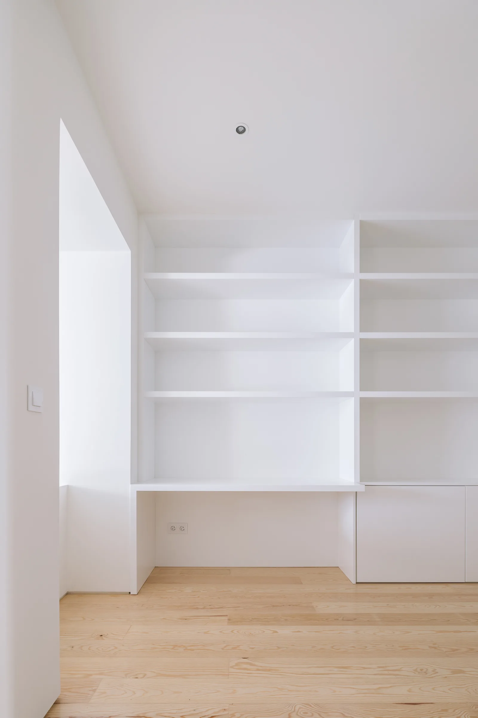 A minimalist room features custom white built-in shelving with an integrated desk space over light wood flooring.