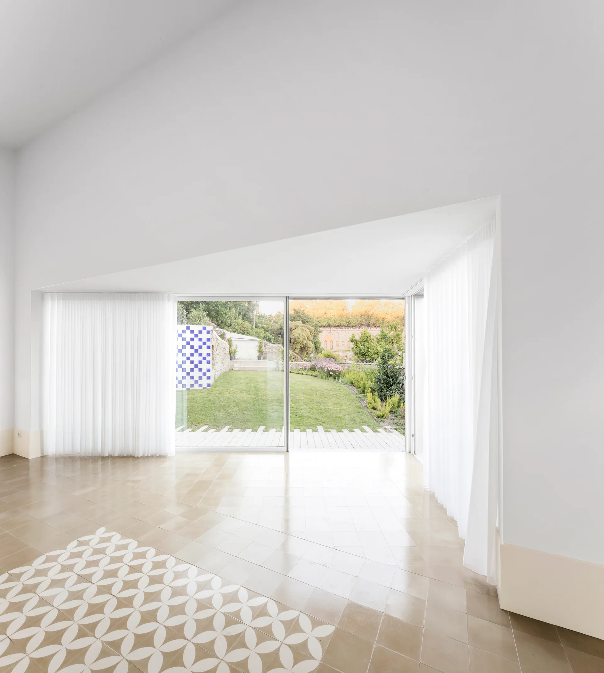 A minimalist room features white walls, a vaulted ceiling, large sliding glass doors opening to a garden, and beige tiled floors with a patterned insert.