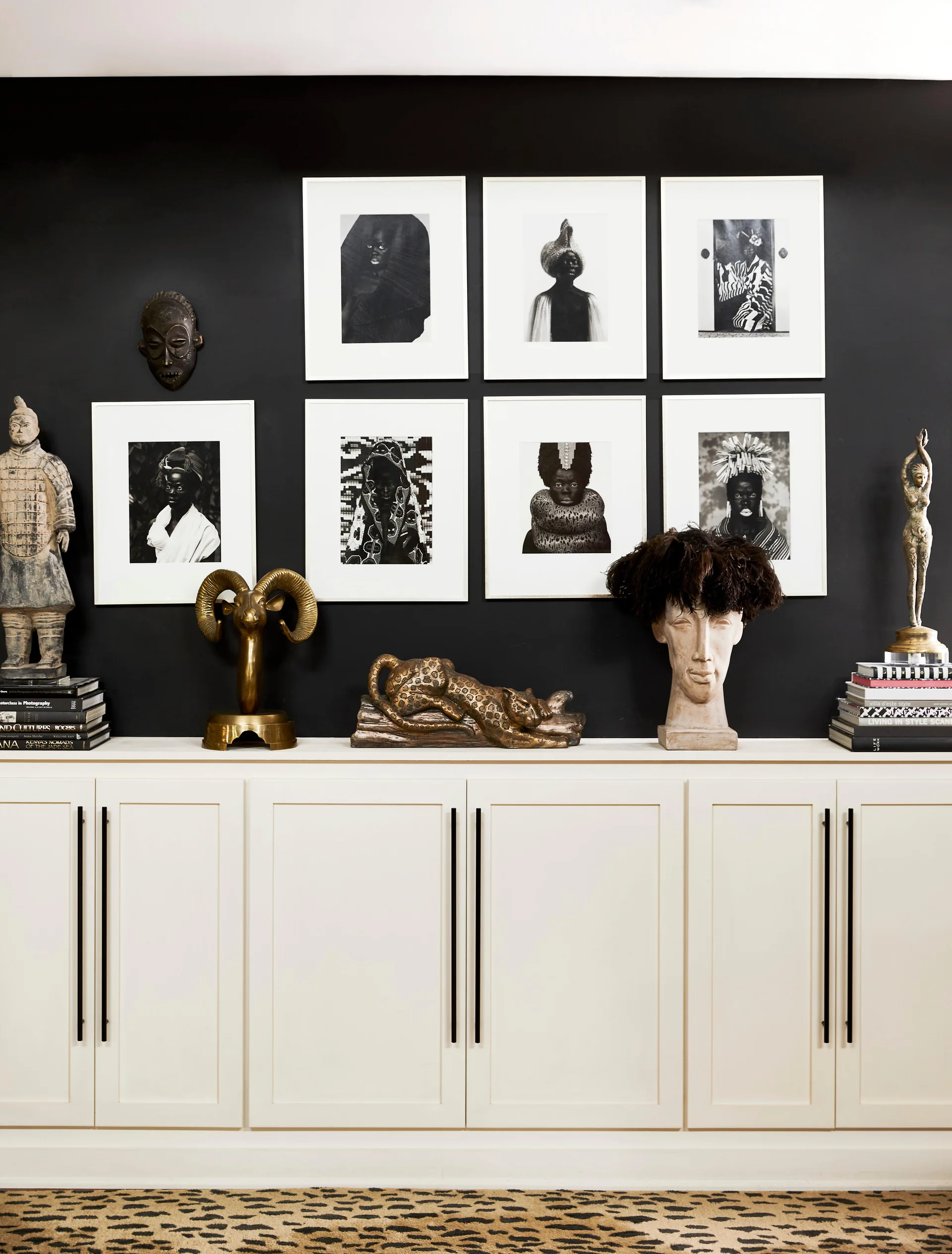 A white built-in cabinet features brass sculptures and books beneath a grid of framed black and white portraits on a matte black wall.