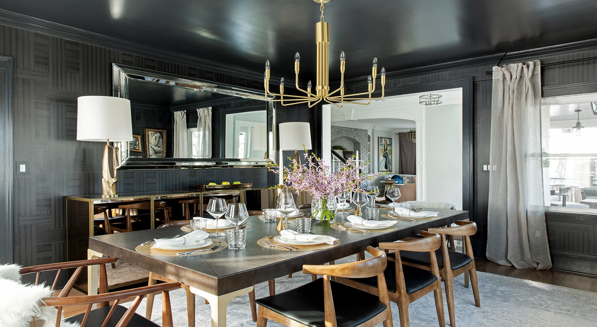 A dark charcoal dining room features a wooden dining table with mid-century chairs under a brass chandelier and a glossy black ceiling.