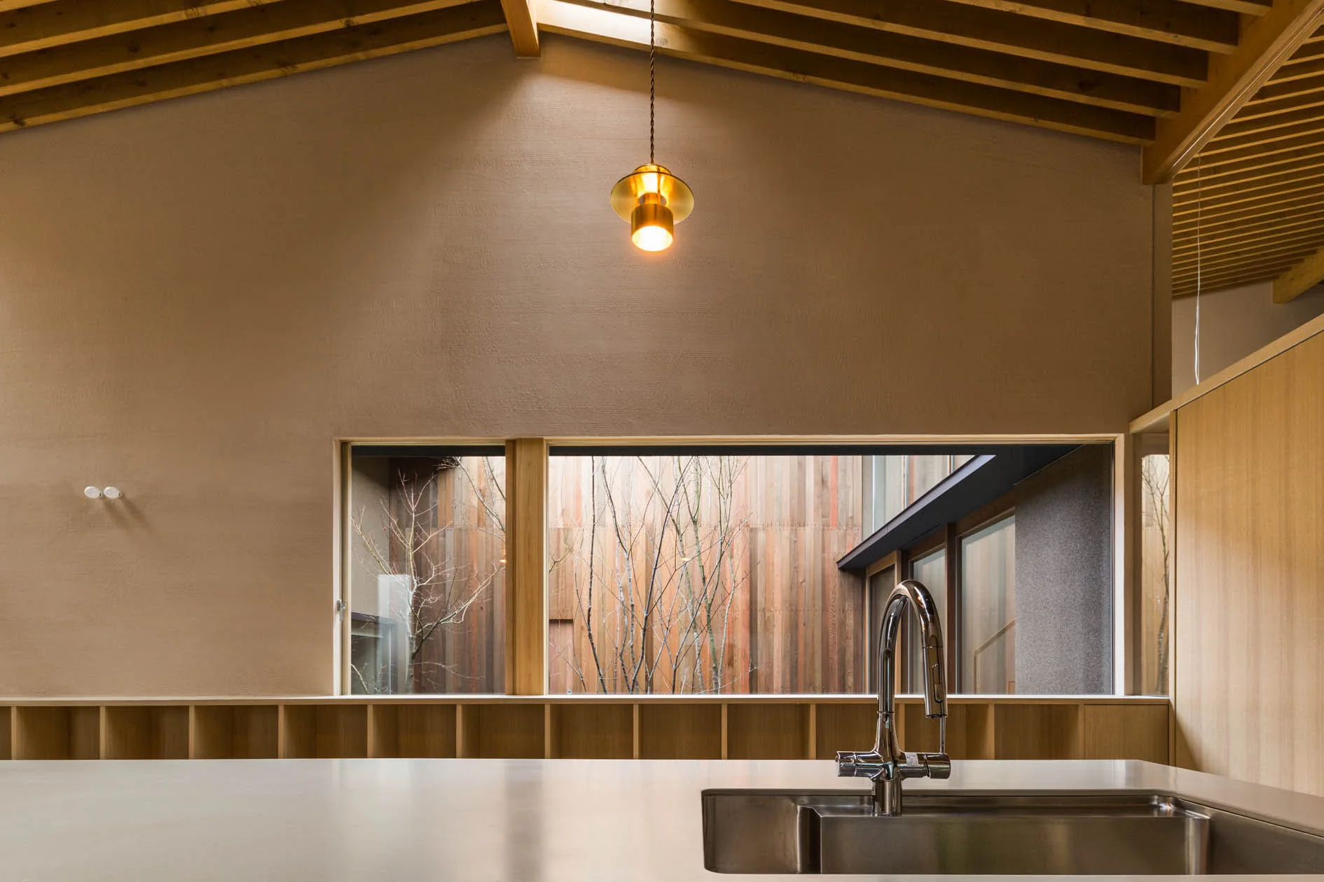 A minimalist kitchen features a stainless steel sink, a long horizontal window looking out to a wood-sided courtyard, and exposed ceiling rafters.