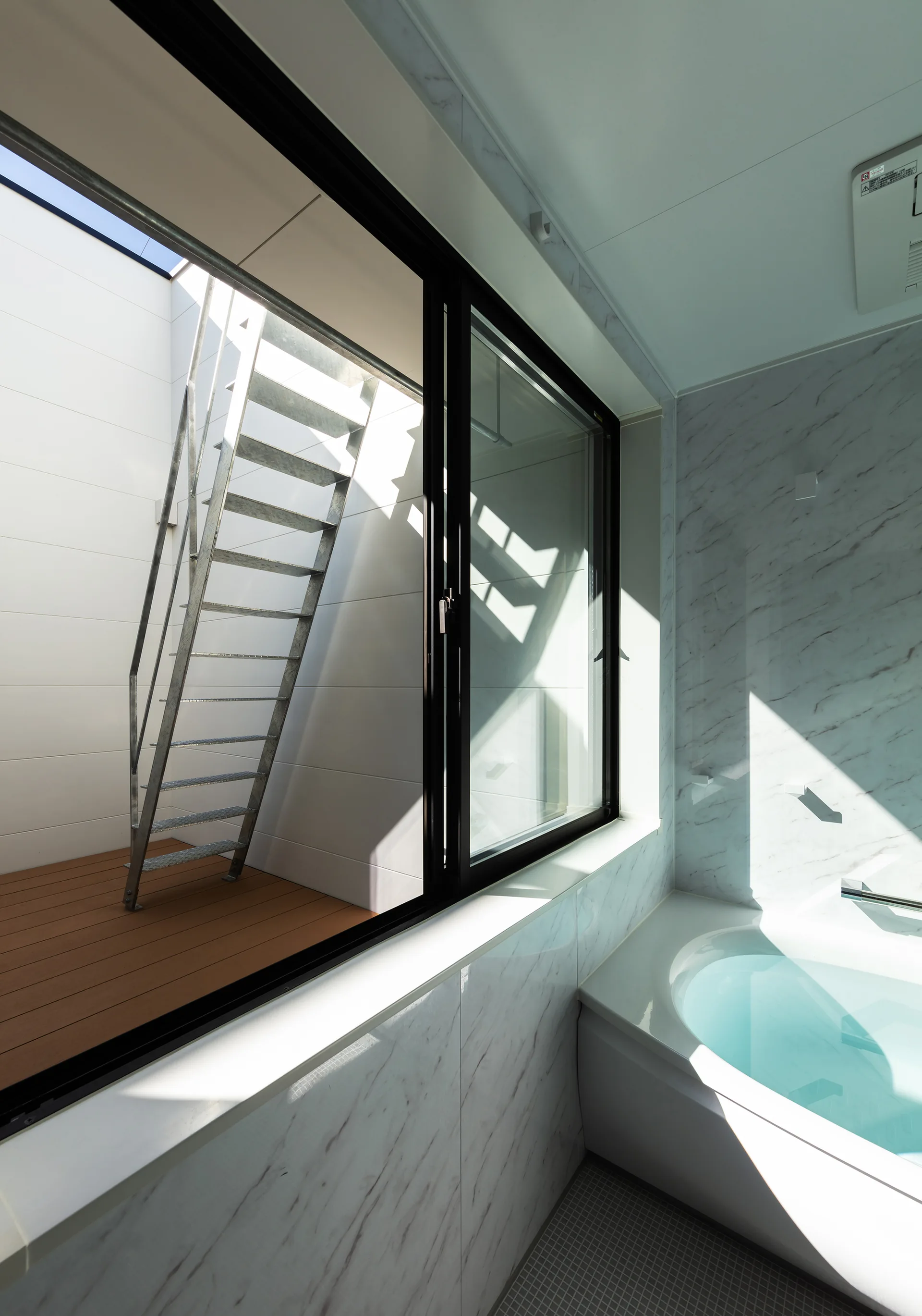 A modern bathroom with a white bathtub and marble tiles features a large black-framed window overlooking a wood deck and metal staircase.