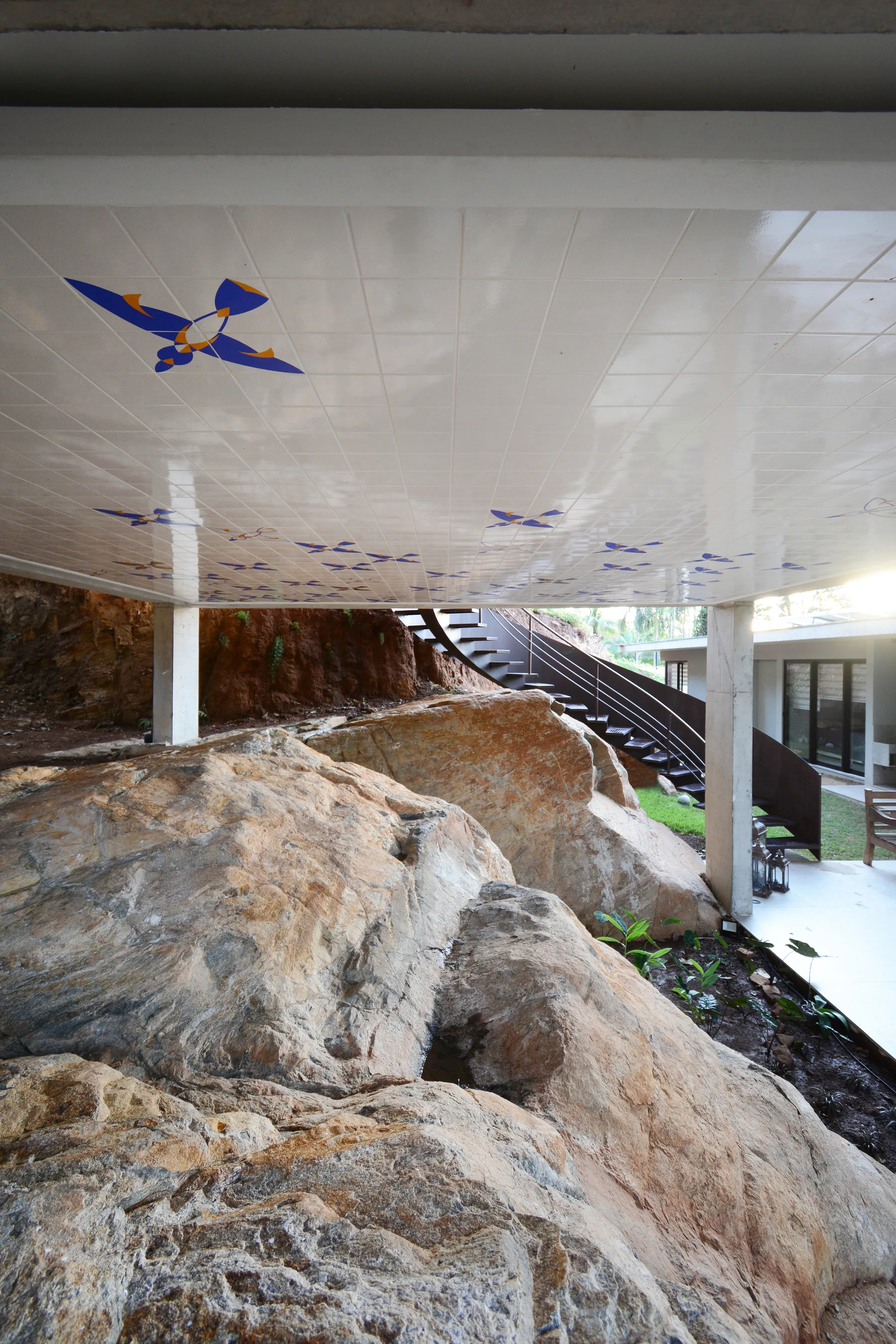 A covered outdoor patio featuring large natural boulders integrated beneath a white tiled ceiling with blue patterns near a metal staircase.