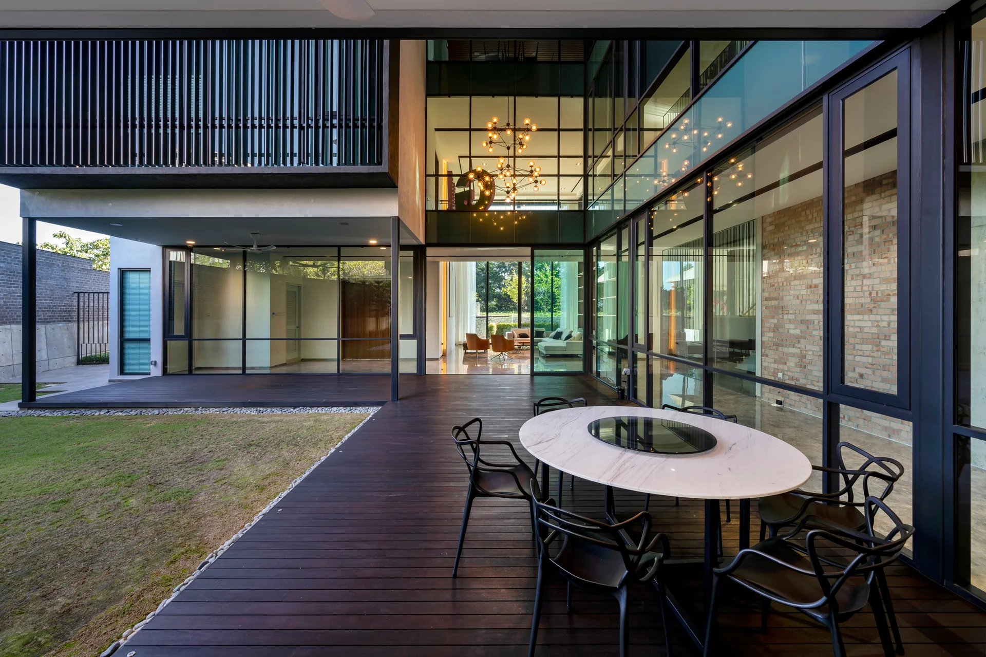 A modern covered dark wood patio deck features an oval white marble dining table with black chairs next to large floor-to-ceiling glass walls.