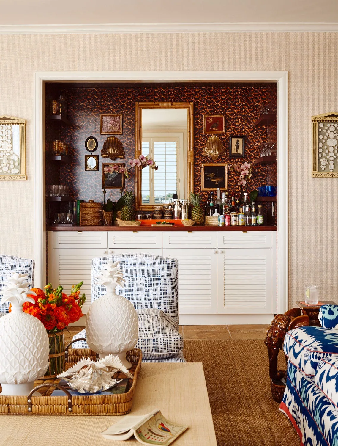 A recessed wet bar featuring white louvered cabinets, tortoiseshell patterned wallpaper, a bamboo-framed mirror, and blue patterned armchairs.