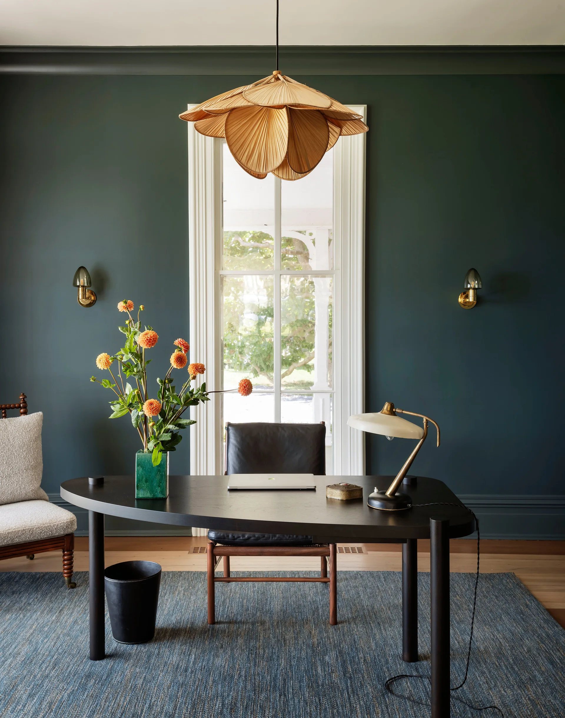 A curved black wood desk with a brass lamp is placed on a blue textured rug against dark green walls and a tall white-trimmed window.