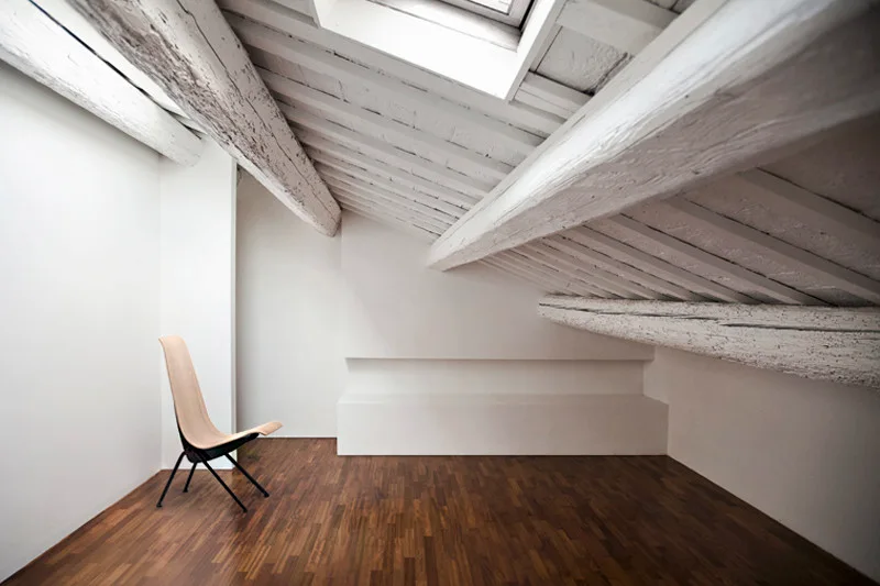 A minimalist attic room with a sloped ceiling, white-painted wood beams, a skylight, dark wood floors, and a single wooden lounge chair.