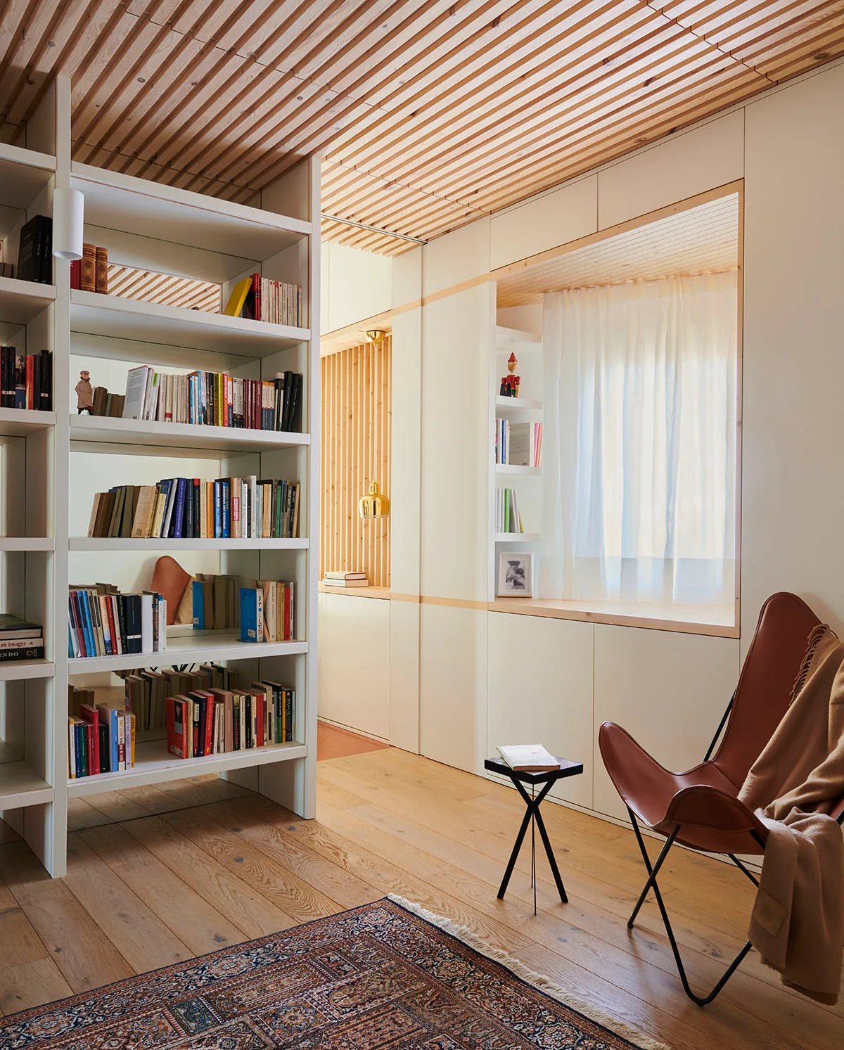 A reading nook features a brown leather butterfly chair, a built-in bookshelf, and a linear wood slatted ceiling over hardwood flooring.