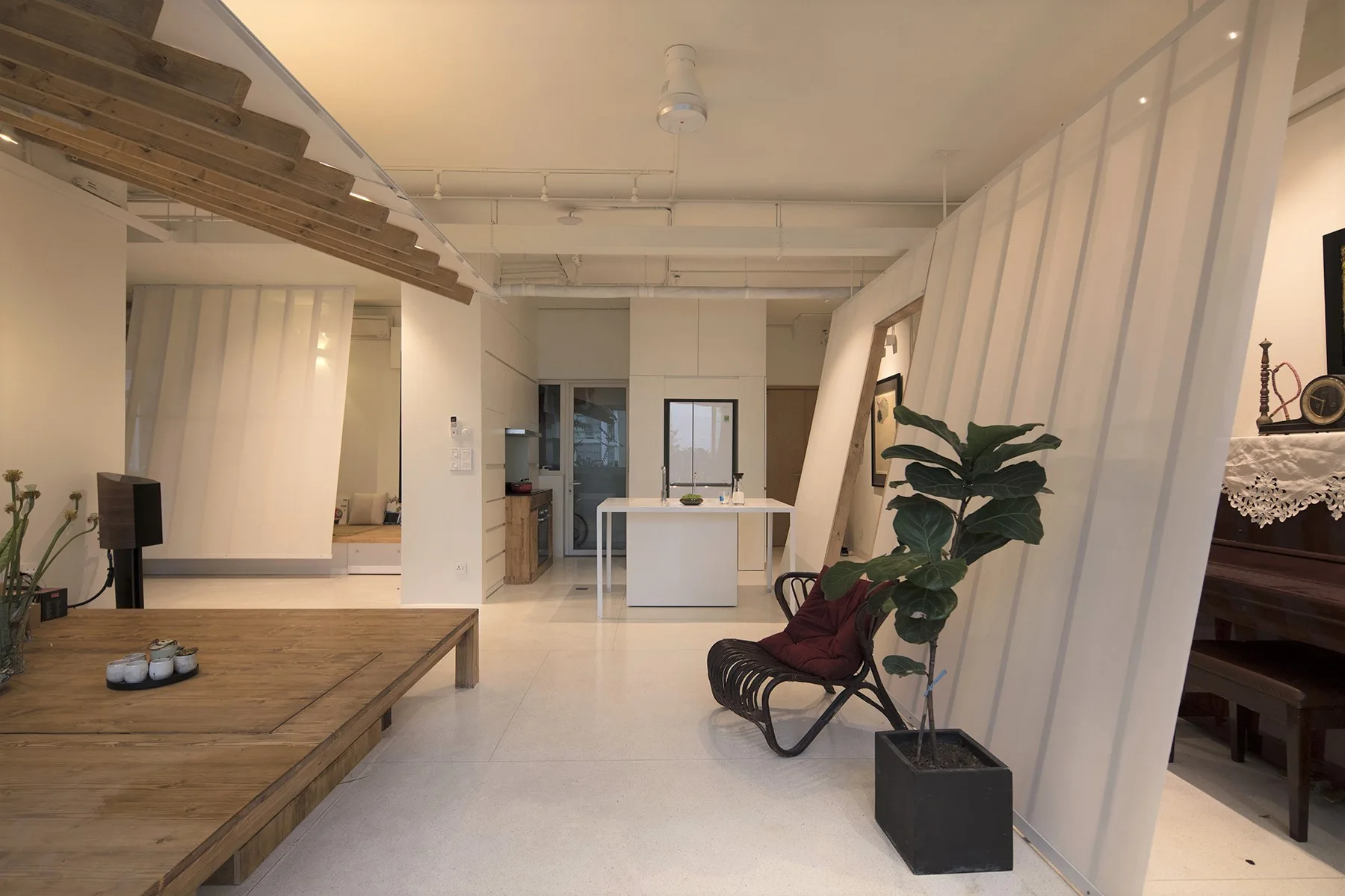 A minimalist open-plan living room featuring slanted white partition walls, a raised wooden platform, and a black accent chair with a potted plant.