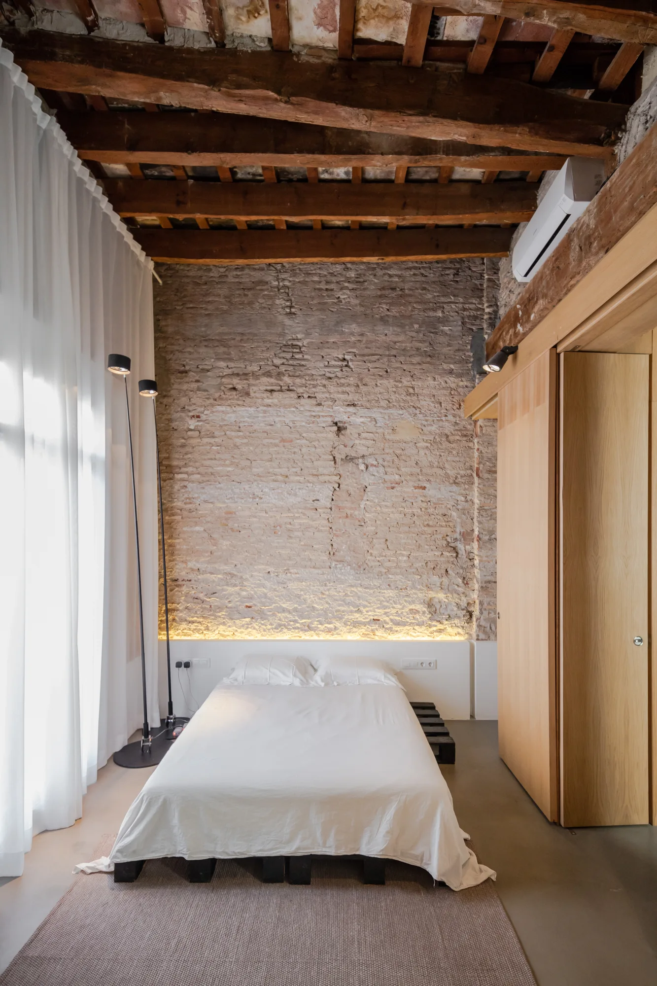 A low bedroom bed sits against an exposed brick wall beneath rustic timber ceiling beams, lit by a warm LED light strip behind the headboard.