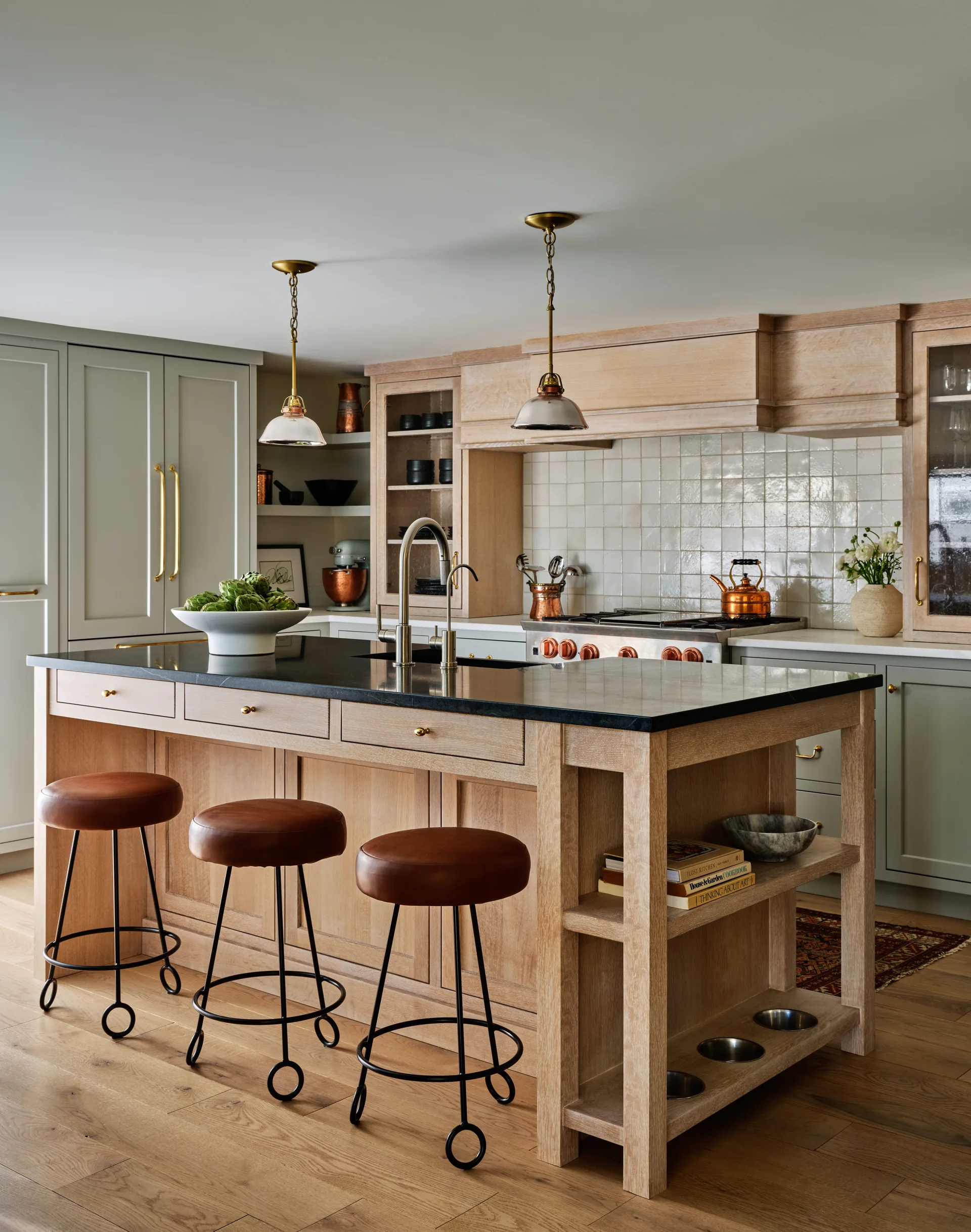 A kitchen featuring a light oak island with leather bar stools, dark countertops, sage green perimeter cabinetry, and brass pendant lights.