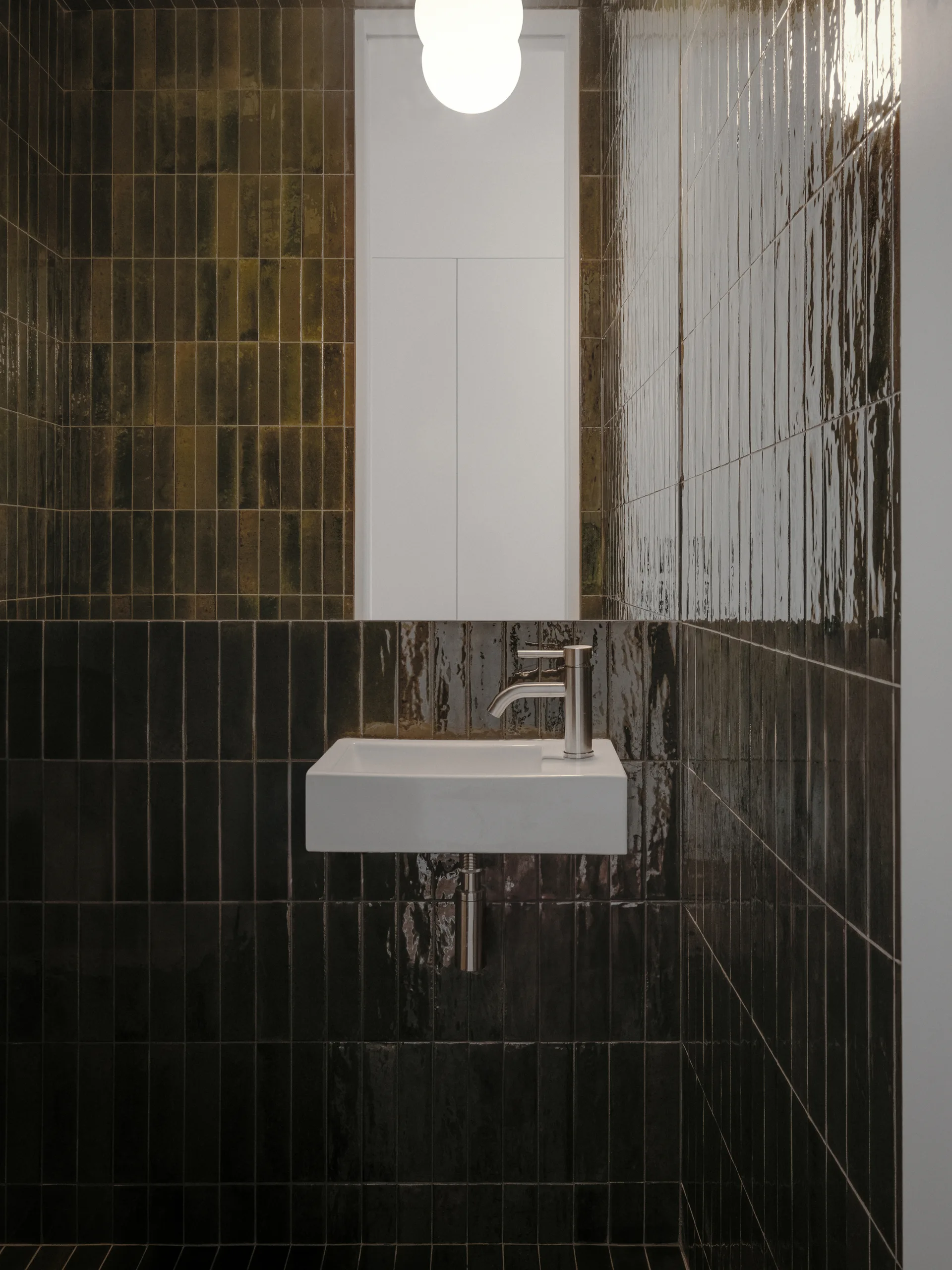 A small powder room features a white wall-mounted sink against dark brown glossy vertical tiles, with a silver faucet and a glowing globe light above.