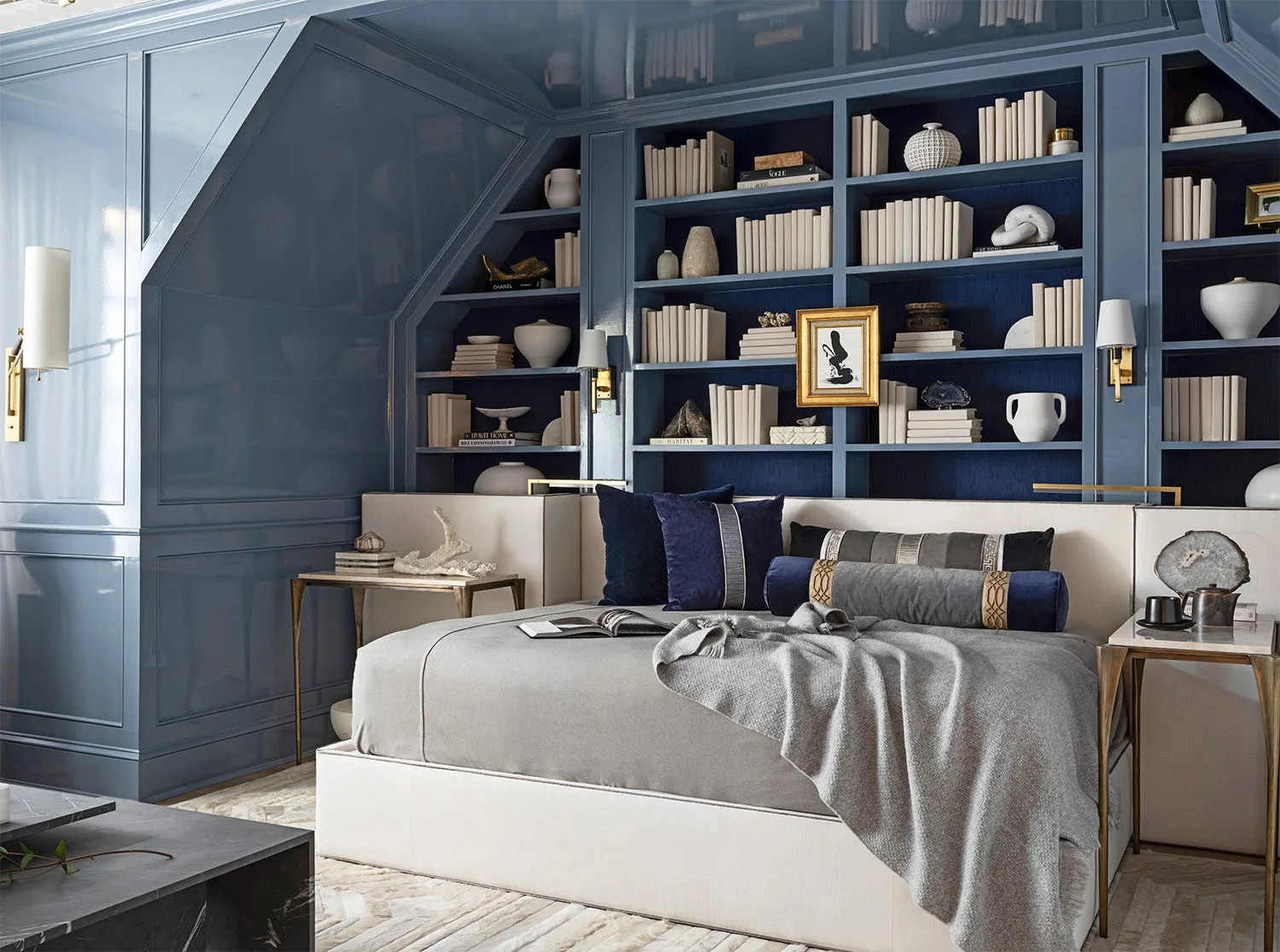 A bedroom featuring blue high-gloss lacquered walls and built-in bookshelves behind a low grey bed with brass wall sconces.