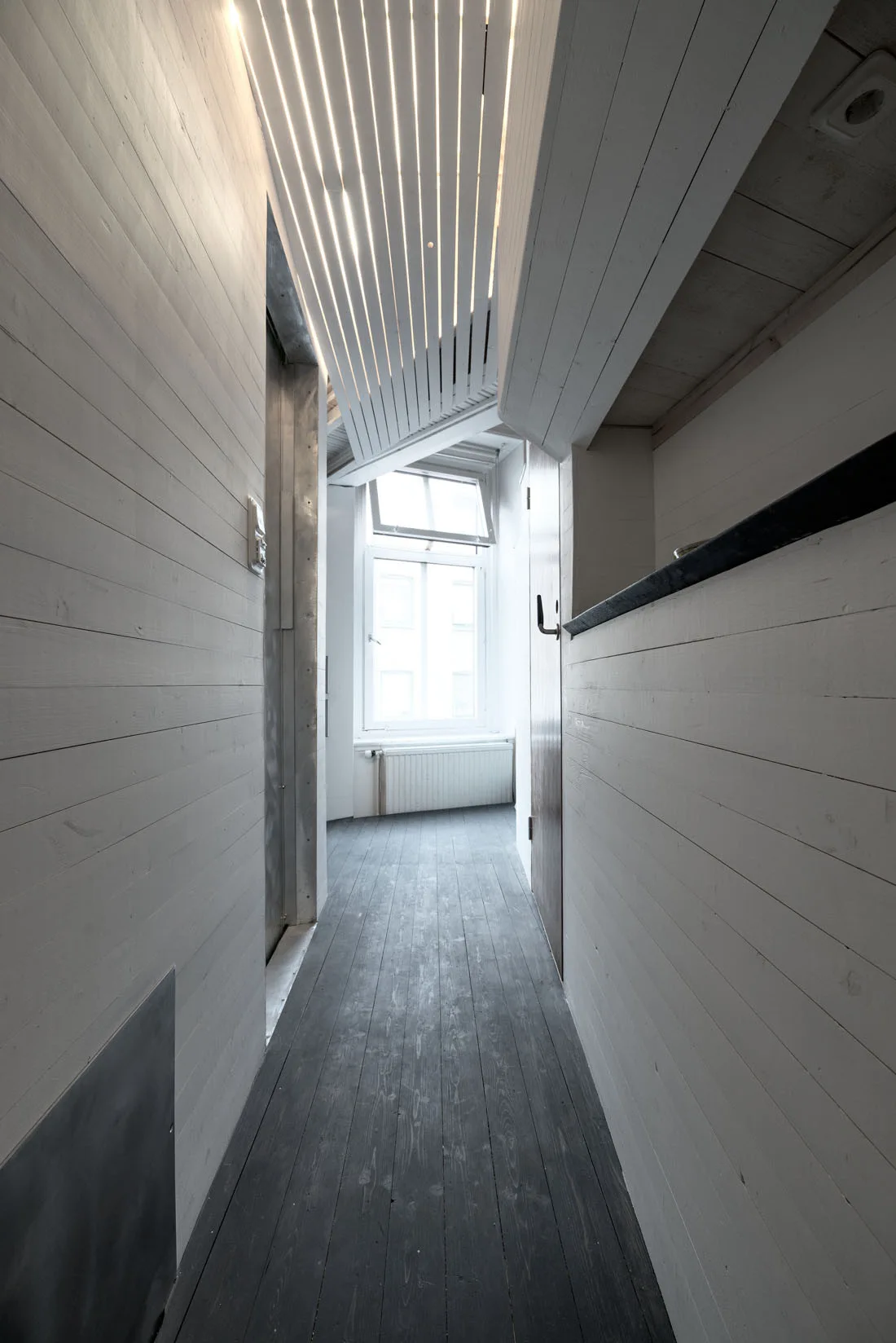 A narrow hallway features white-painted wood paneled walls, a dark wood plank floor, and a geometric slatted ceiling with integrated linear lighting.