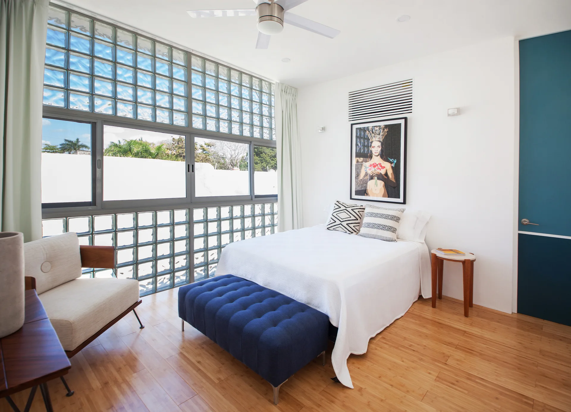A modern bedroom features a bed with white linens, a tufted blue bench, an armchair, and a large wall of glass blocks.