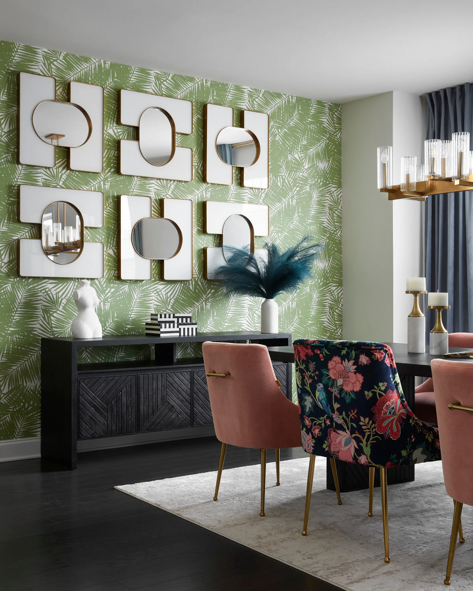 A dining room features green palm-leaf wallpaper, a black wooden sideboard, six modern mirrors, pink velvet chairs, and a floral upholstered chair.
