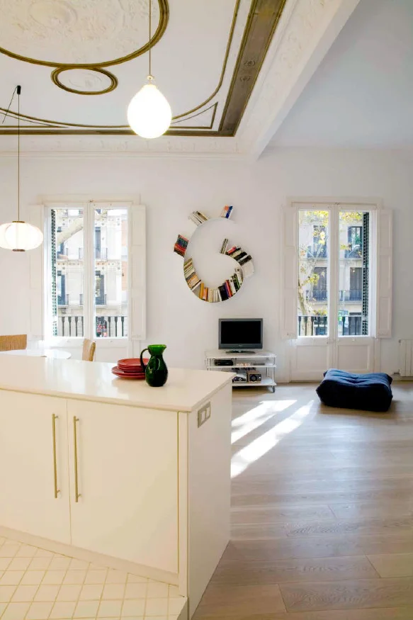 A bright open-concept room features a white kitchen island, a spiral-shaped wall bookshelf, and two windows with white shutters under an ornate ceiling.
