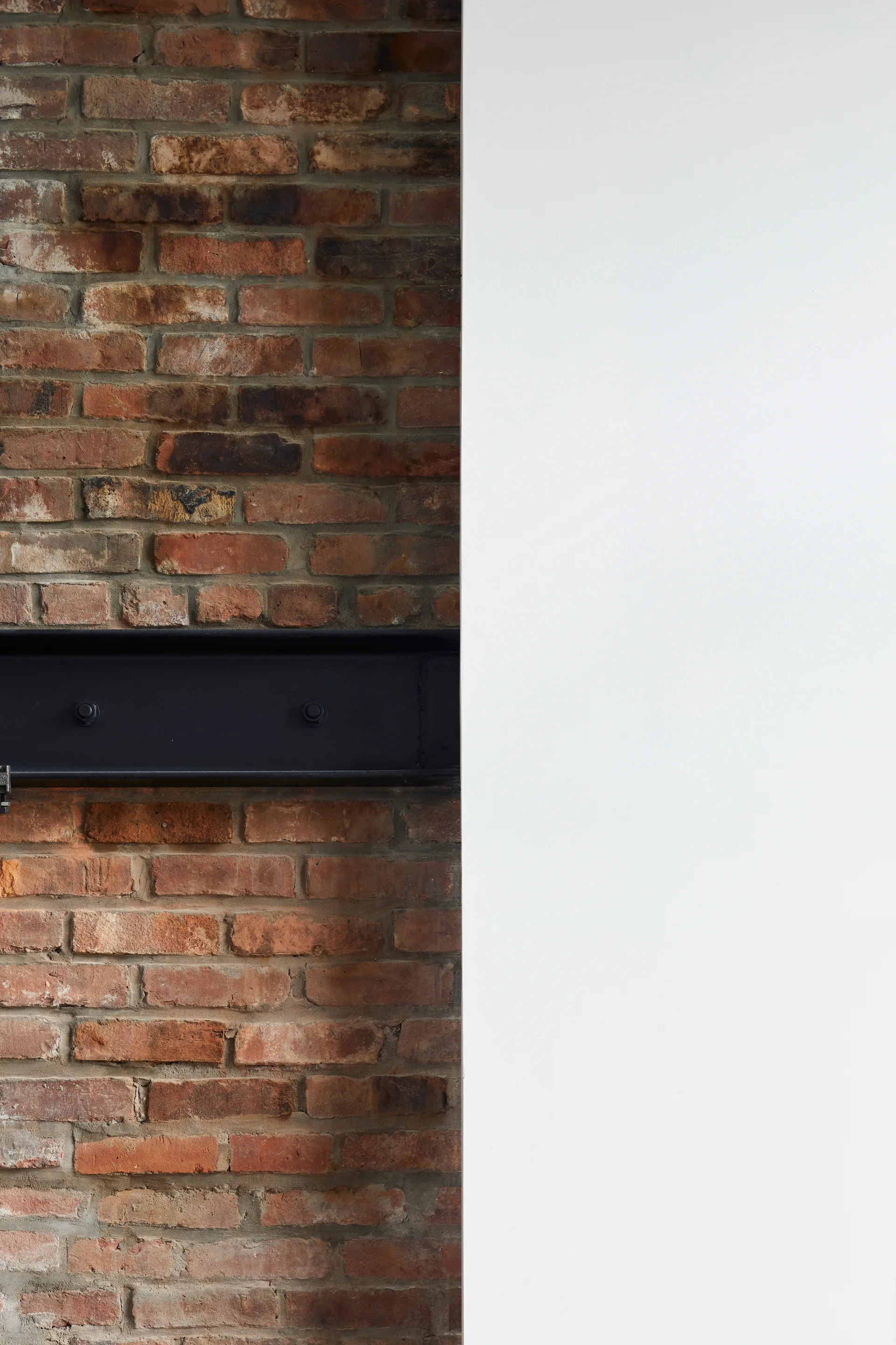 A horizontal black structural steel beam is set against an exposed red brick wall next to a smooth white plastered wall.