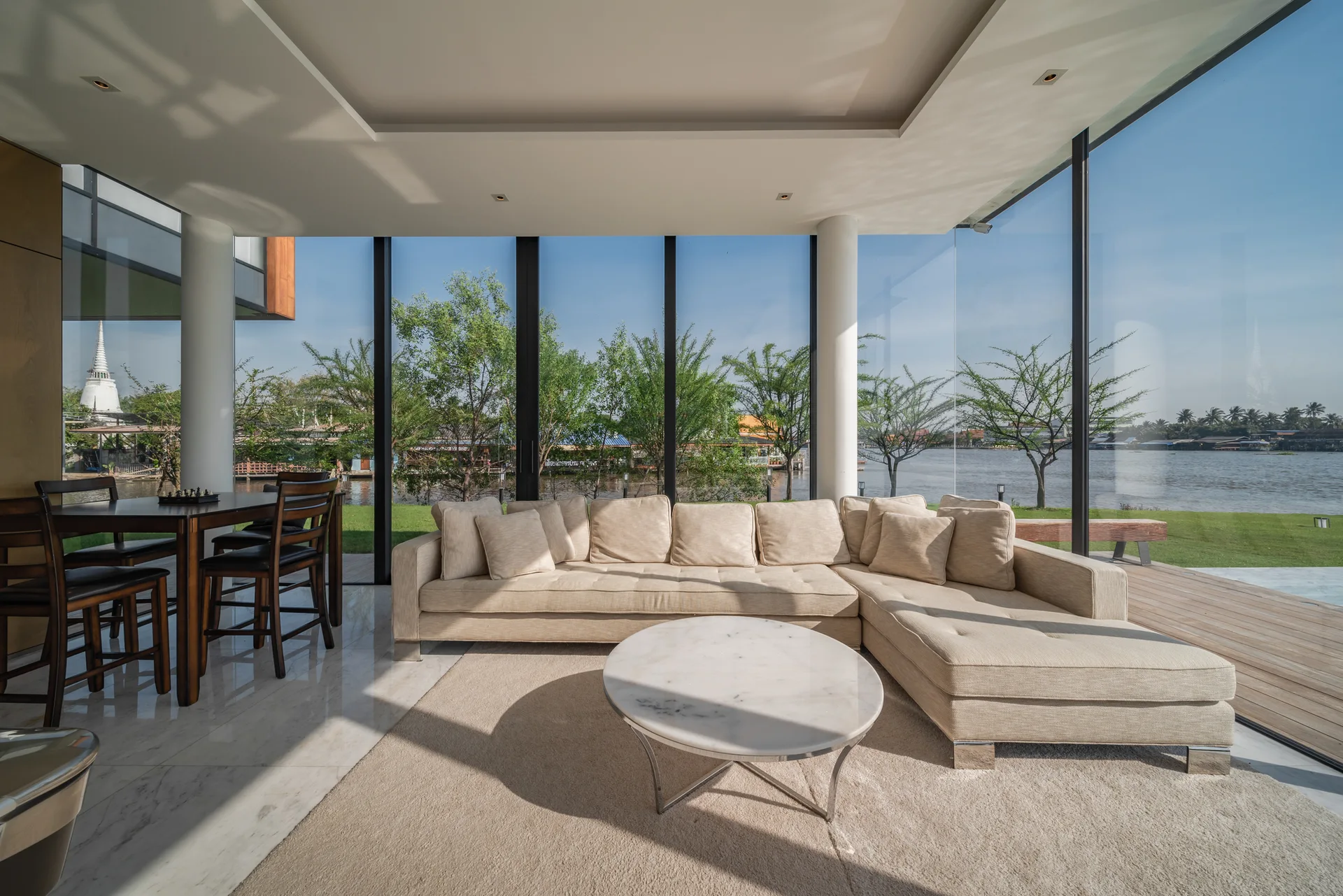A modern riverfront living room with a beige sectional sofa, a round marble coffee table, and large glass walls overlooking a river.