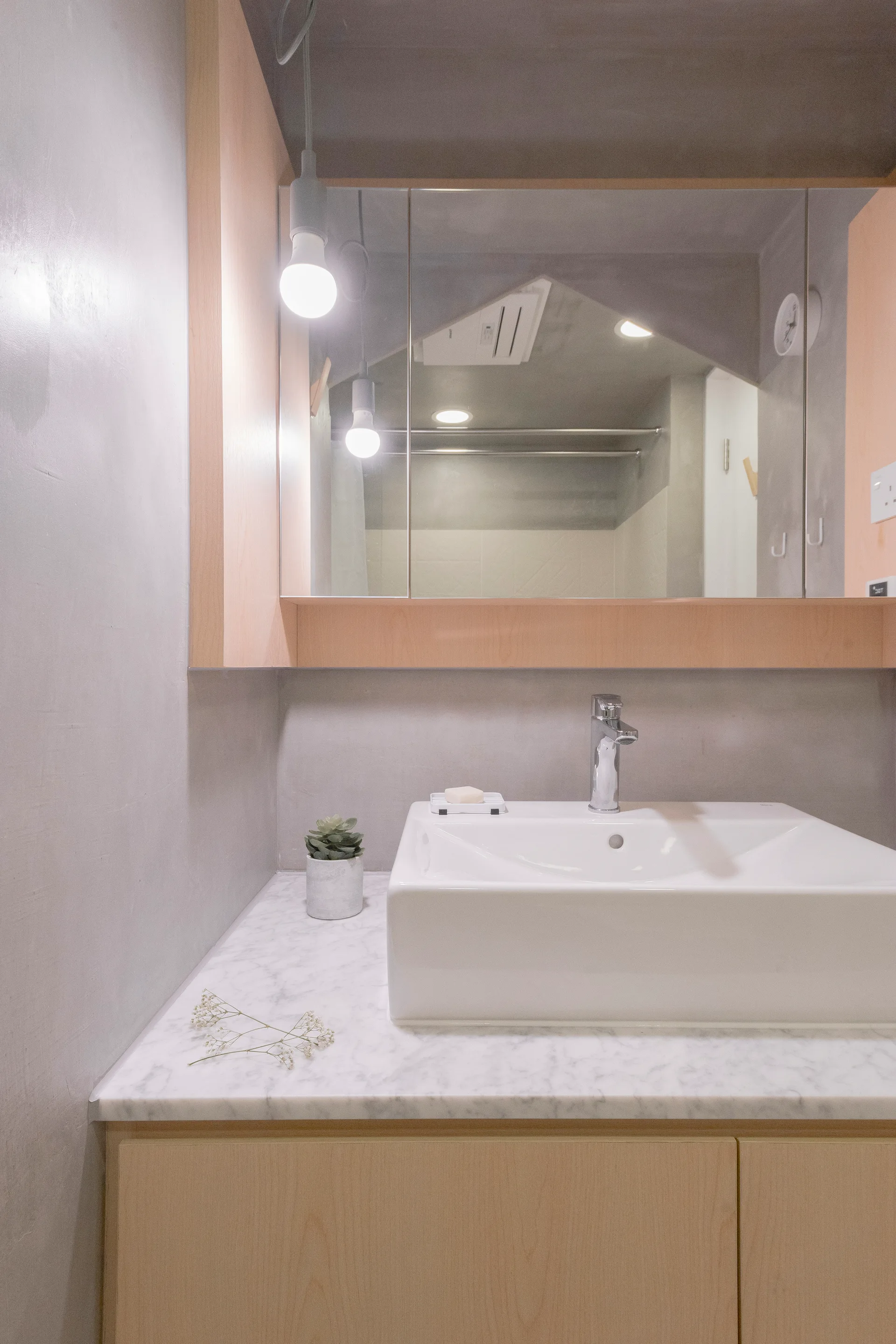 A modern bathroom vanity features a white ceramic sink, a marble countertop, light wood cabinetry, and a mirrored cabinet against grey plaster walls.