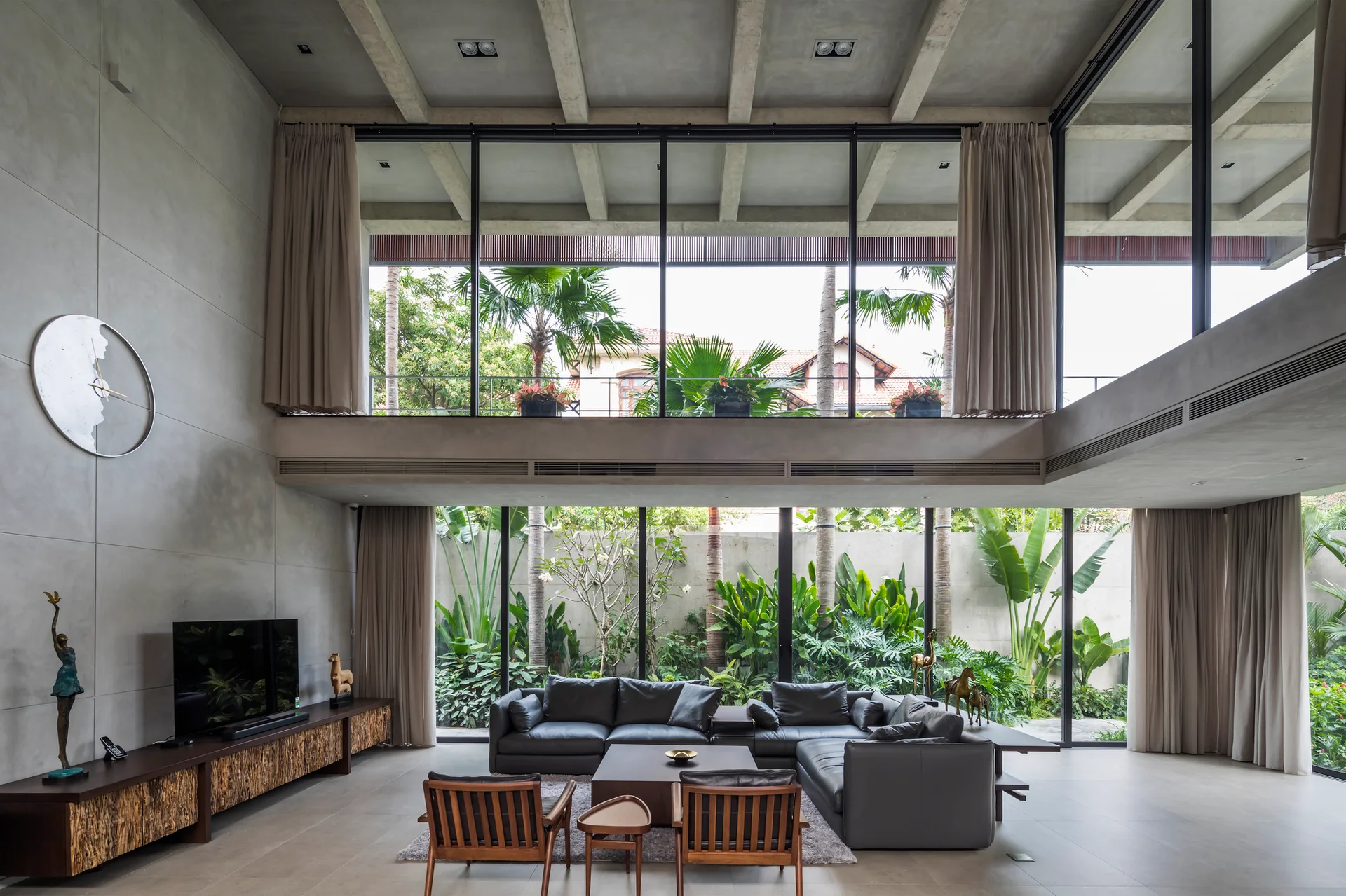 A spacious double-height living room features exposed concrete beams, a grey sectional sofa, and floor-to-ceiling windows showcasing a lush green garden.