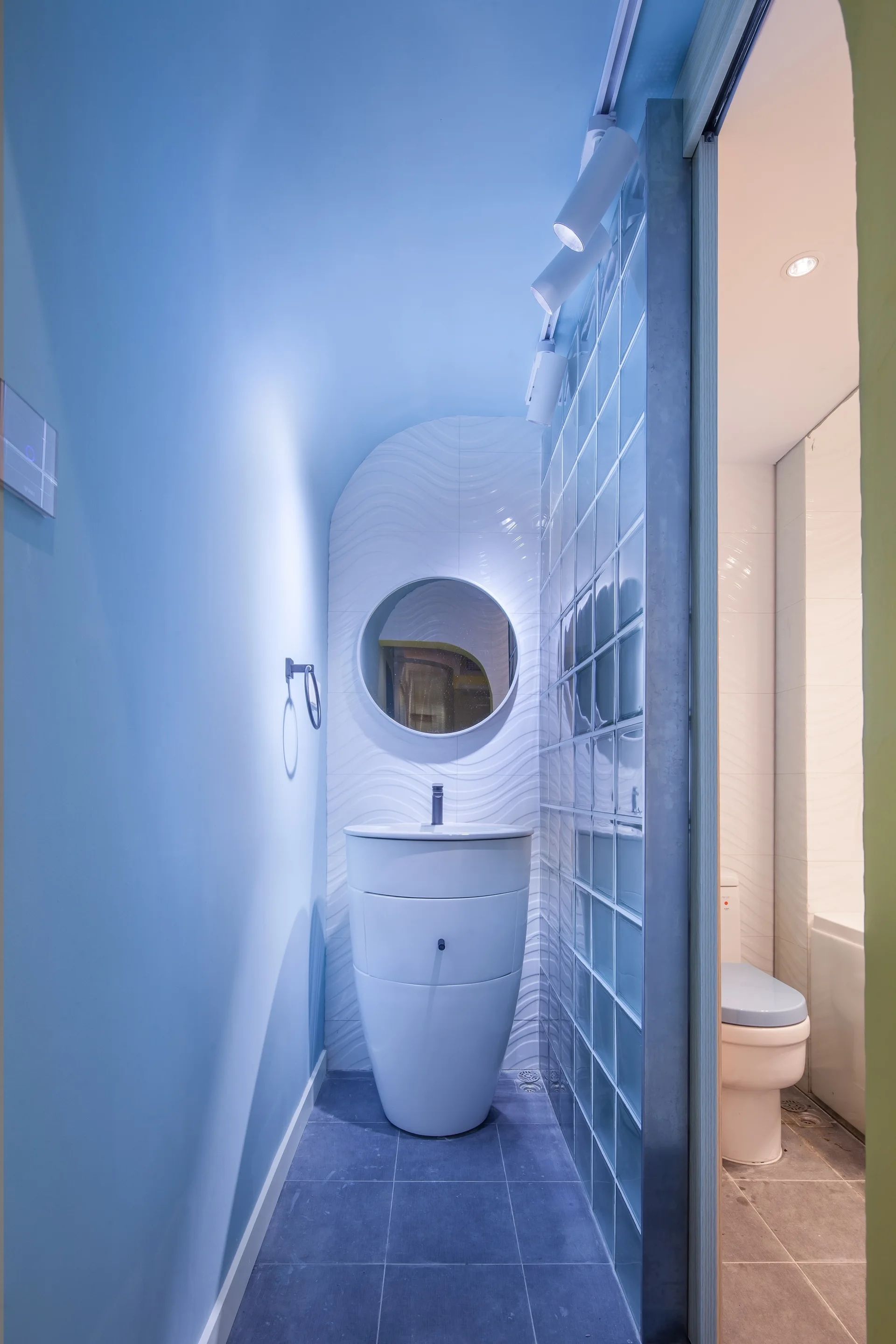 A compact blue bathroom featuring a curved white pedestal sink, a round mirror, and a translucent glass block partition wall.