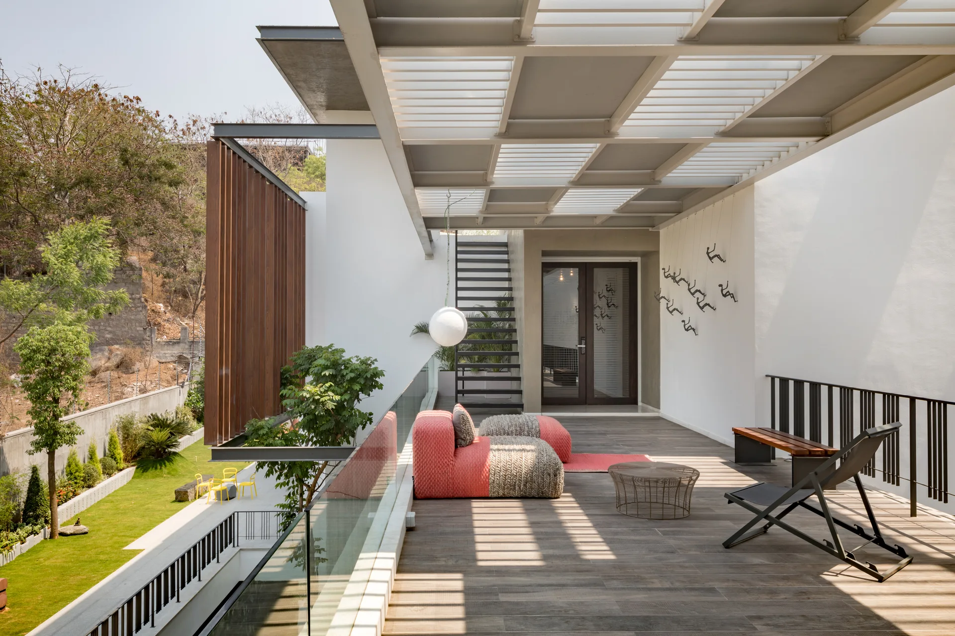 A modern outdoor terrace features a metal louvered pergola, a low-profile pink and grey sofa, wood-patterned flooring, and a glass railing.