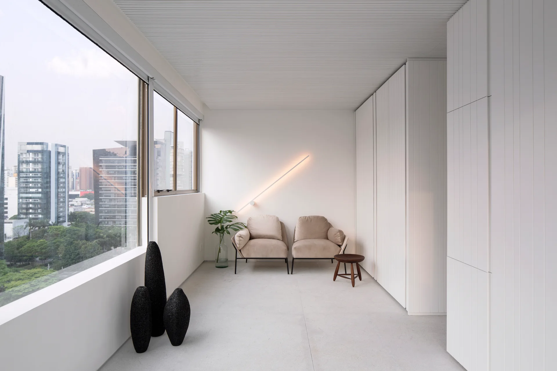 A minimalist room features two beige armchairs below a diagonal wall light, with white built-in cabinets and a large window overlooking a city.