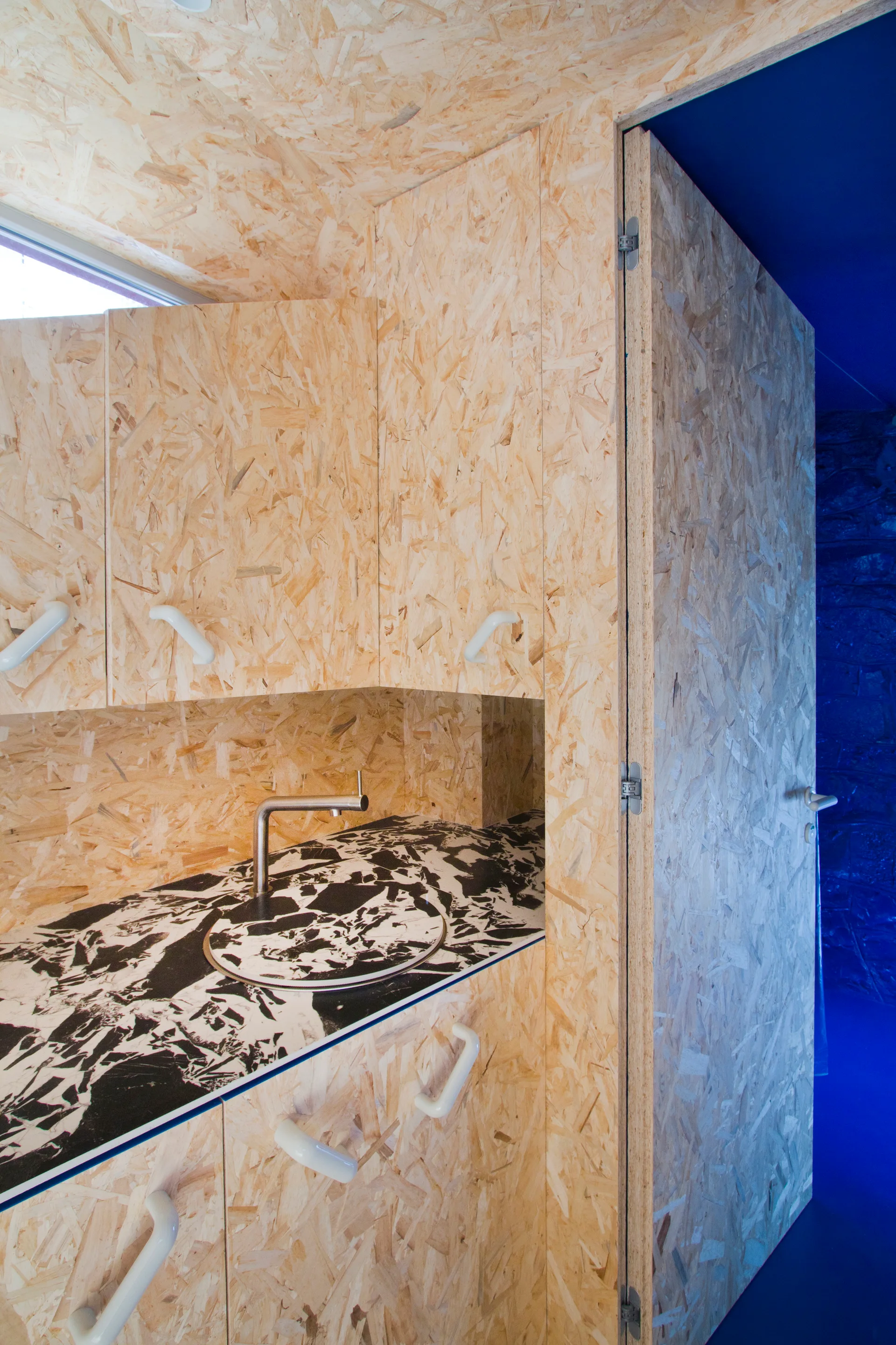A compact kitchen features cabinets clad in oriented strand board, a black and white patterned countertop with a round sink, and white handles.