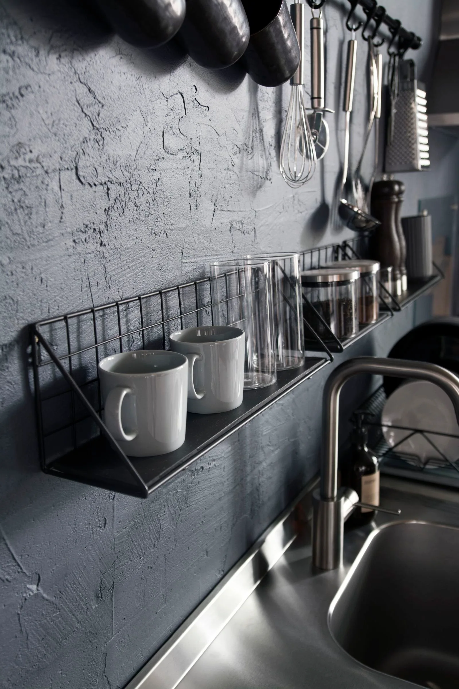 A black wire shelf with white mugs and glass tumblers hangs on a dark textured kitchen wall above a stainless steel sink.
