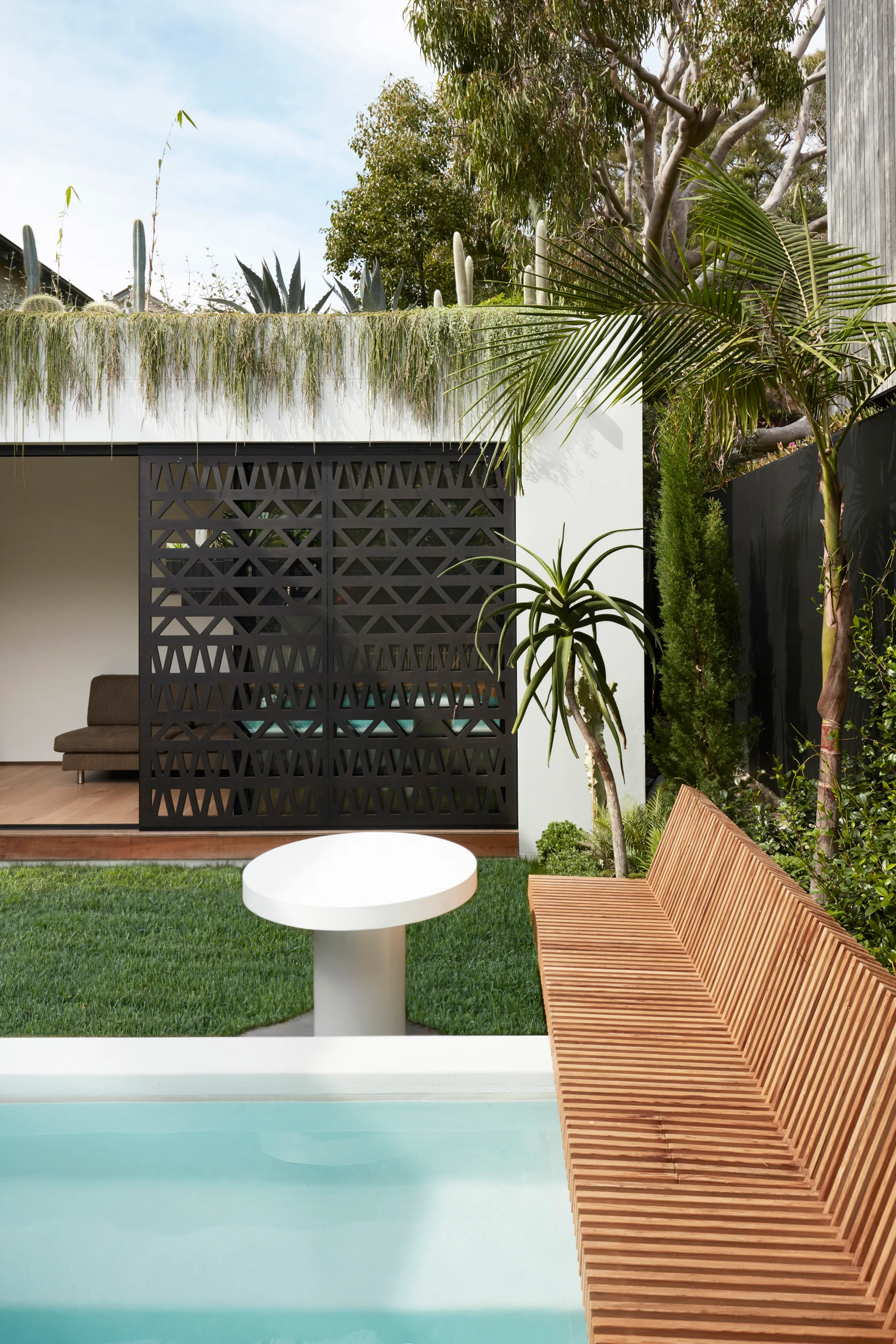 An outdoor pool area with a built-in wooden slatted bench, a white round pedestal table on the lawn, and a black geometric sliding screen door.