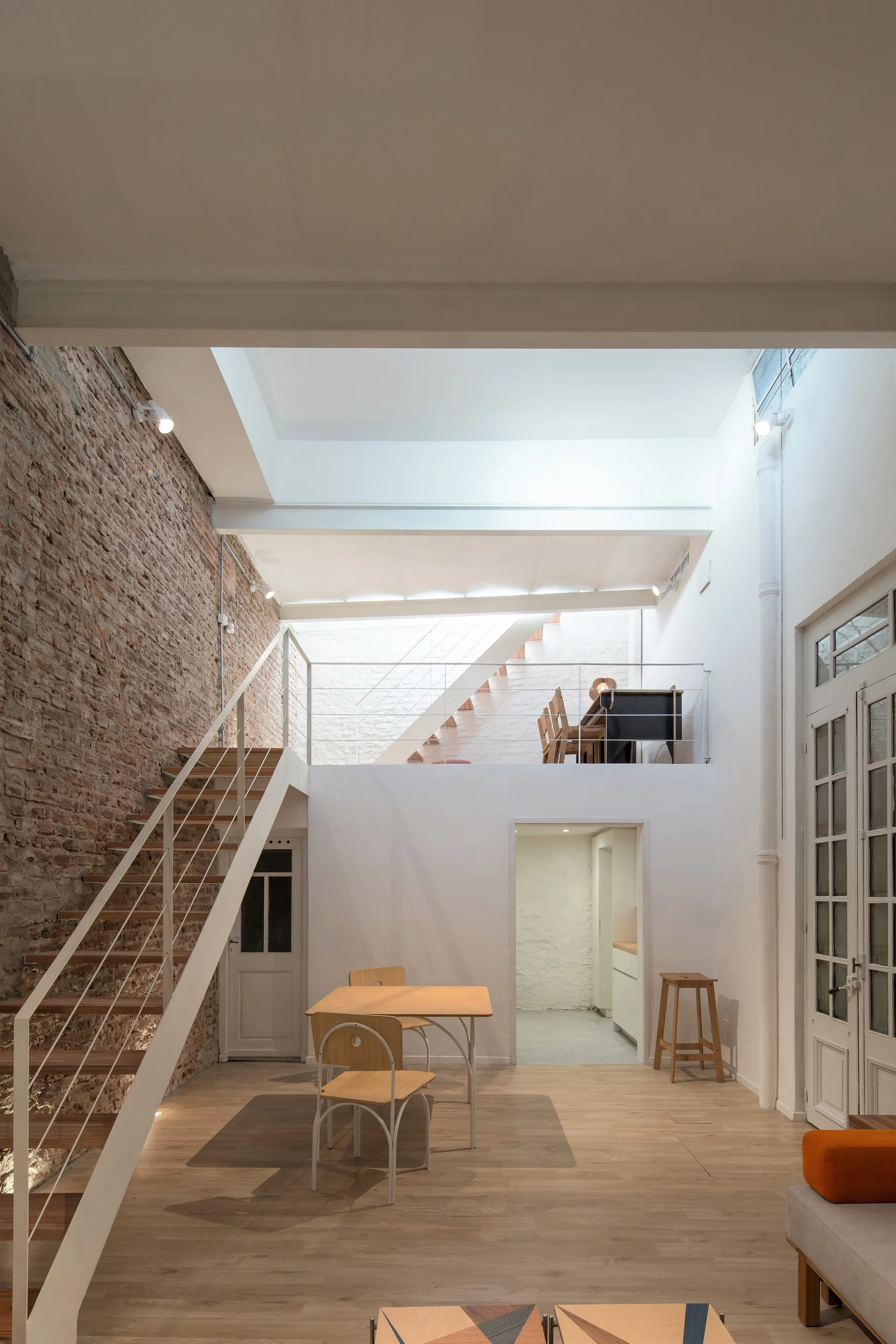 A split-level loft apartment features an exposed brick wall, a white metal open-riser staircase, light wood floors, and a mezzanine level.